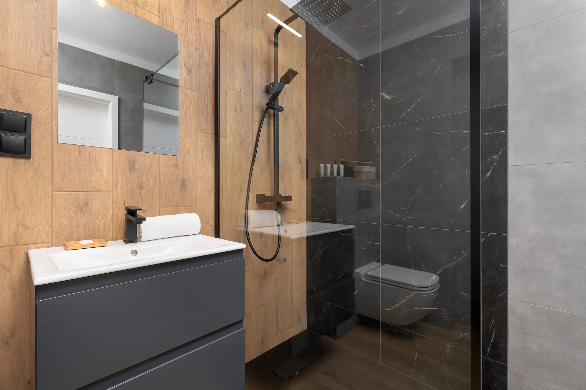 A functional bathroom features a wall-mounted wooden vanity unit with a white sink and a large mirror. A glass-enclosed shower stall with a black rainfall showerhead is visible on the right.