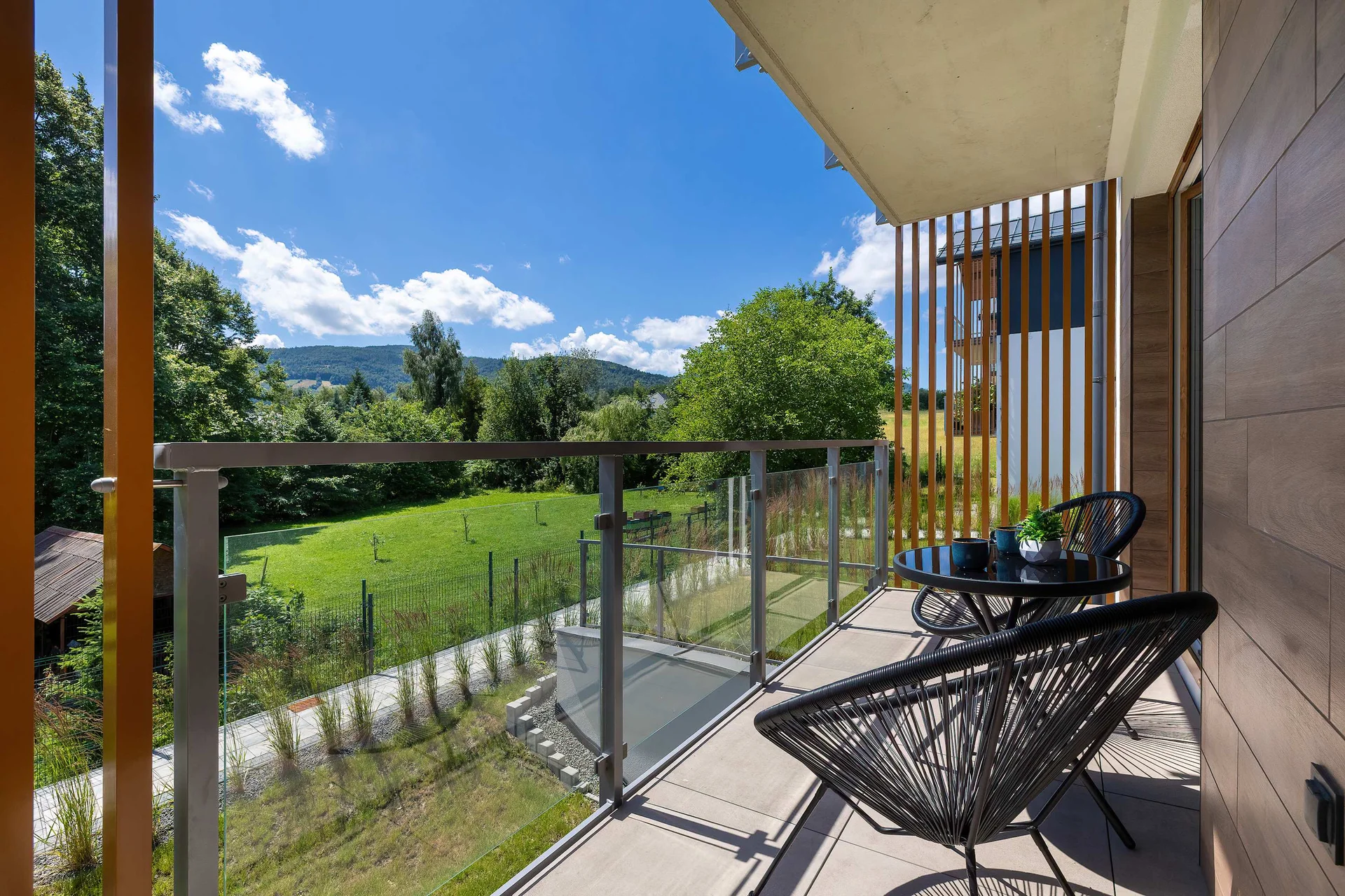 An outdoor balcony is equipped with a black mesh chair and a small round table. The railing overlooks a lush green field with rolling hills visible under a clear blue sky.