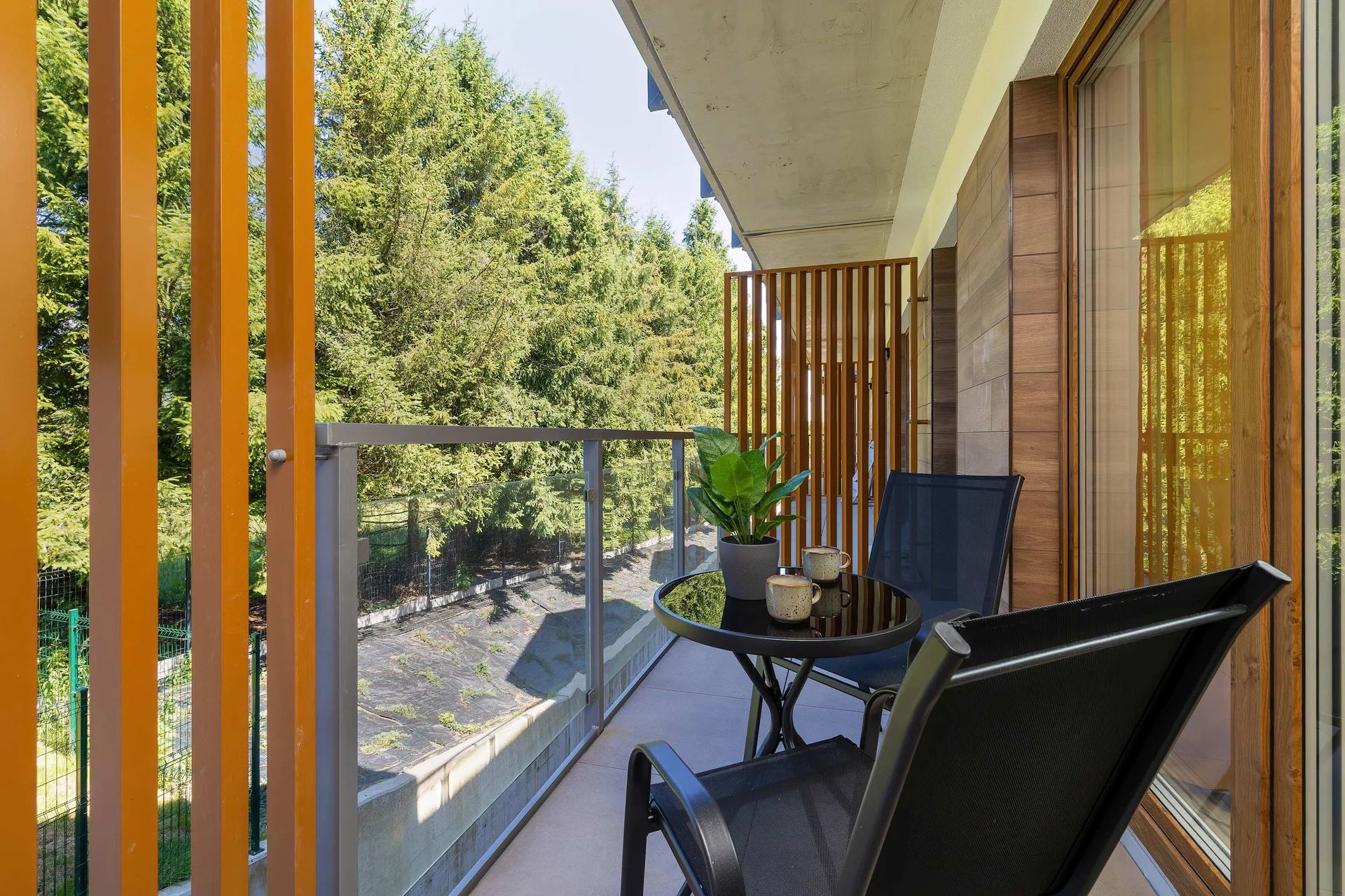 An outdoor balcony is equipped with a black mesh chair and a small round table. Wooden slatted privacy screens enclose the balcony area, which overlooks surrounding trees.