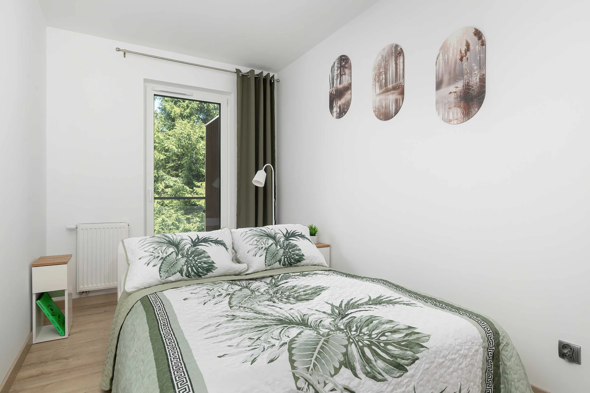 A neat bedroom contains a double bed dressed in white linens with a green floral and leaf pattern. The plain white wall behind the bed creates a clean, minimalist look.