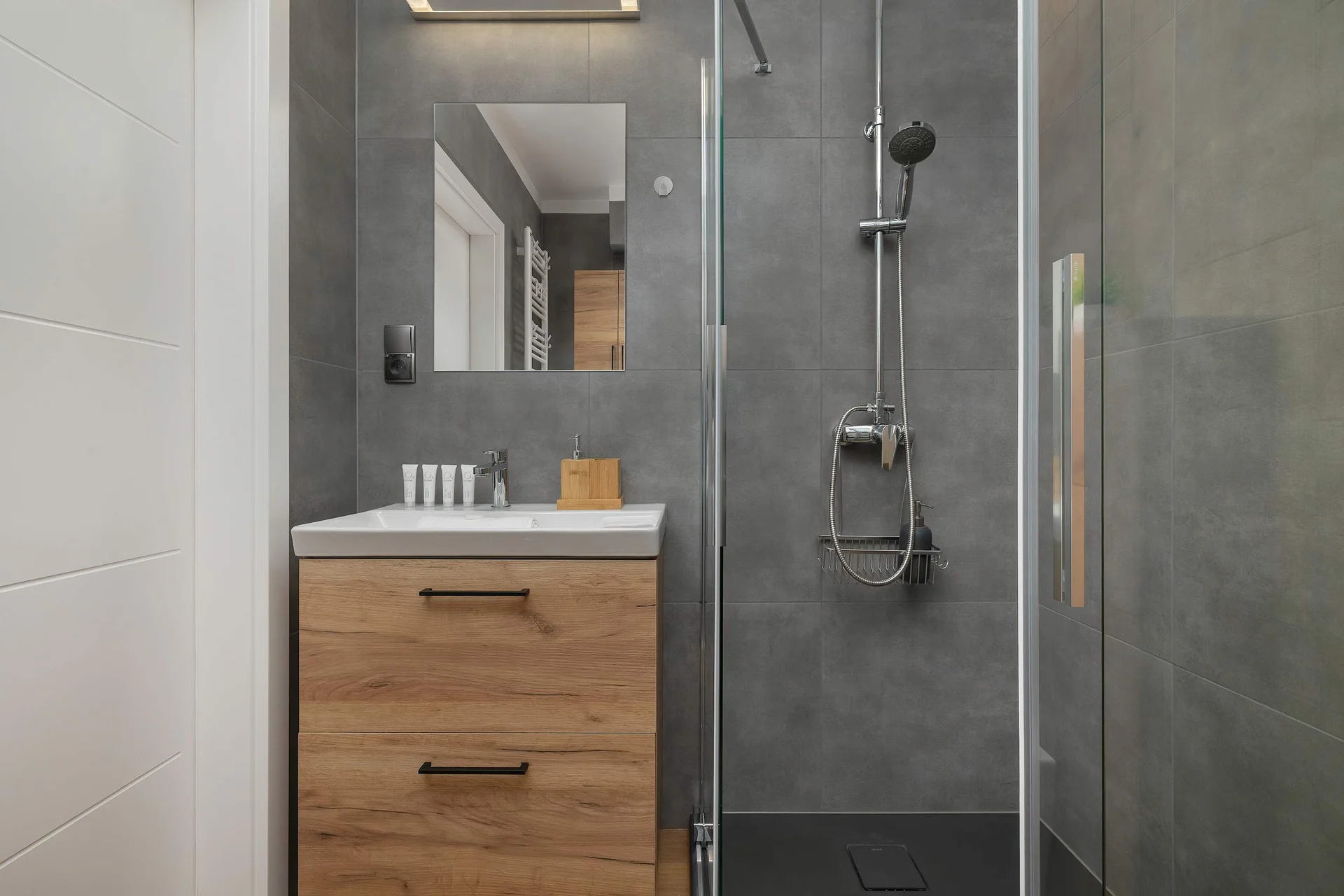 A functional bathroom features a wall-mounted wooden vanity unit with a white sink. A glass-enclosed shower stall with a rainfall showerhead is visible on the right.