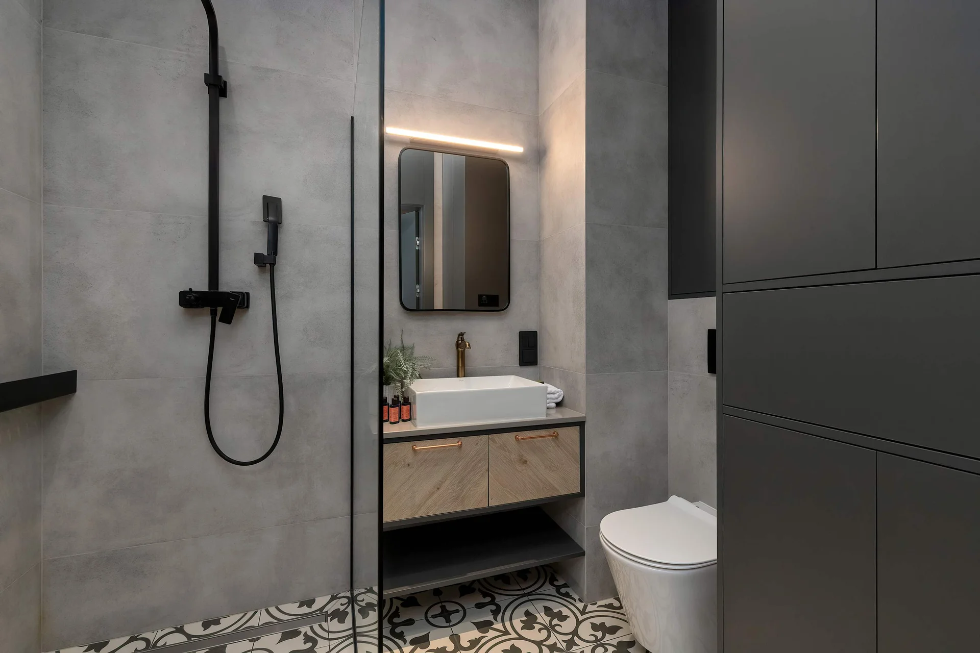 A contemporary bathroom combines smooth concrete-look grey wall tiles with a vibrant terrazzo floor. A dark washbasin vanity sits near a wall-hung toilet.