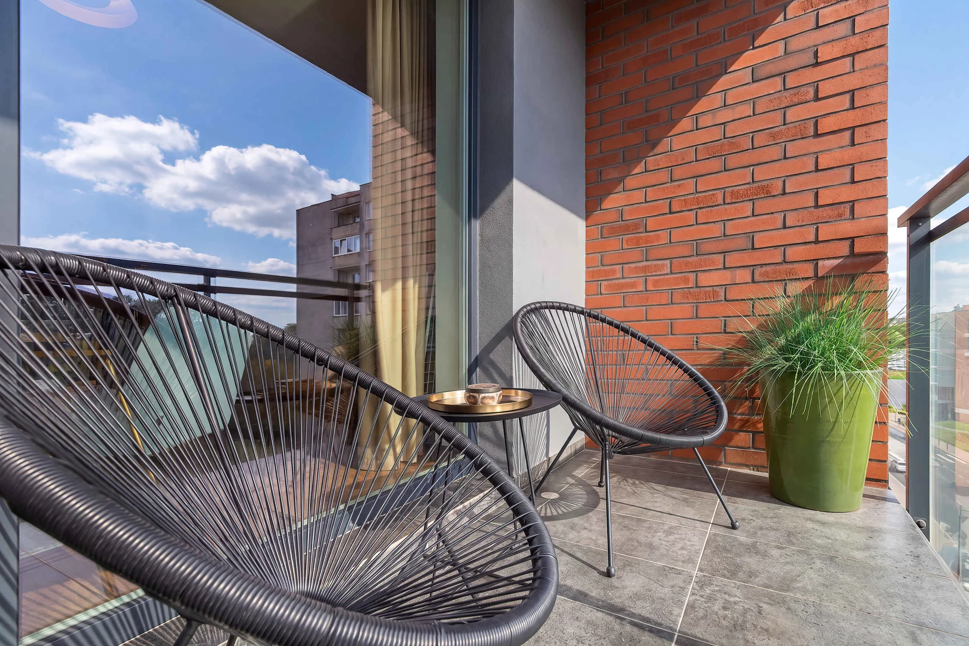 An outdoor balcony with brick accents is furnished with two black string egg chairs and a small table. A green planter sits in the corner under a blue sky.