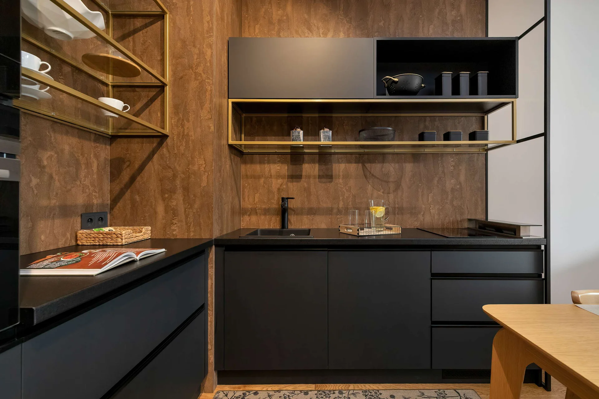 A sleek minimalist kitchen boasts matte black cabinetry paired with warm wood-grained walls. Open brass shelves hold glasses and kitchenware.