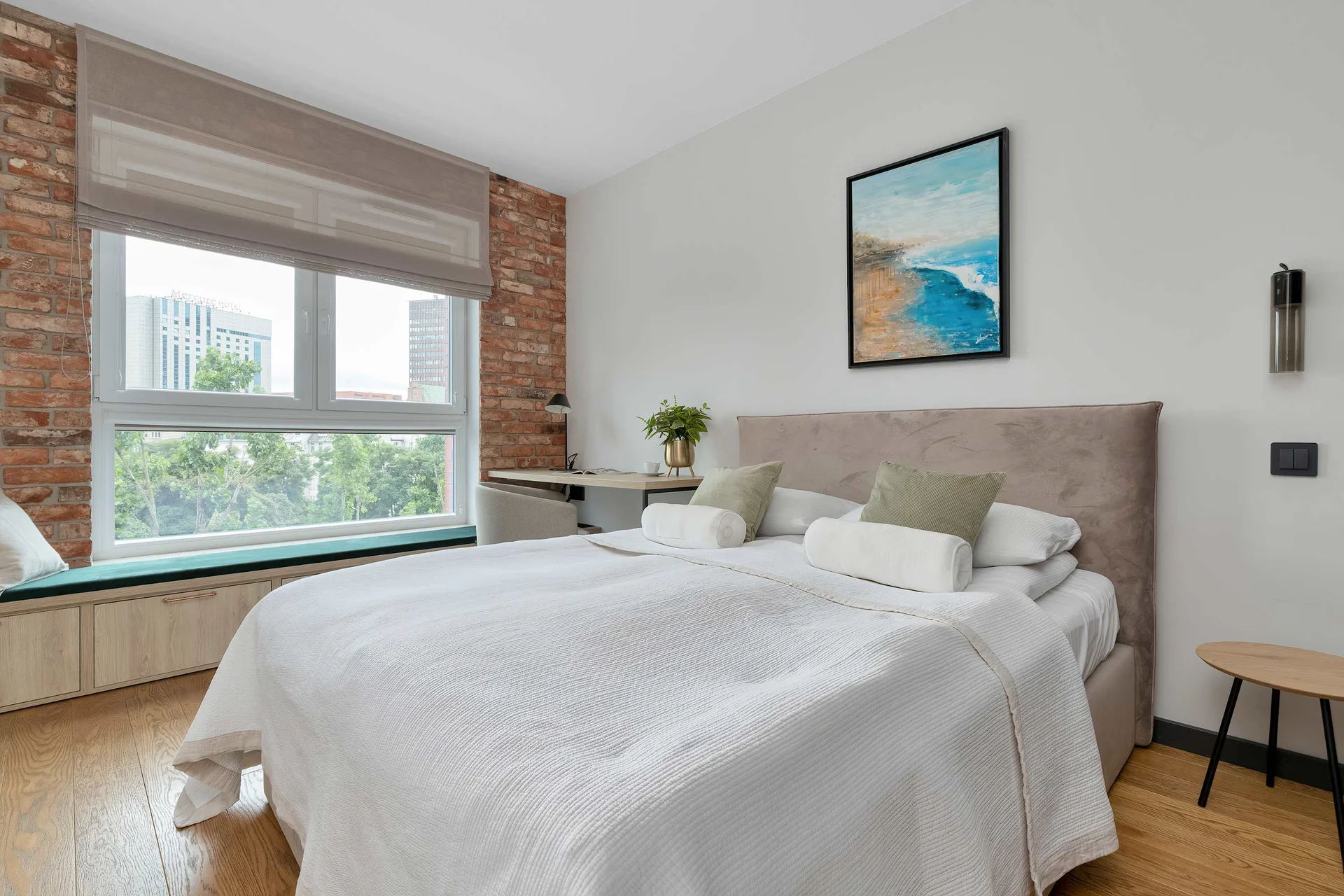 The bedroom showcases a double bed with a light grey tufted headboard and crisp white linens. An industrial brick accent wall frames the large window.