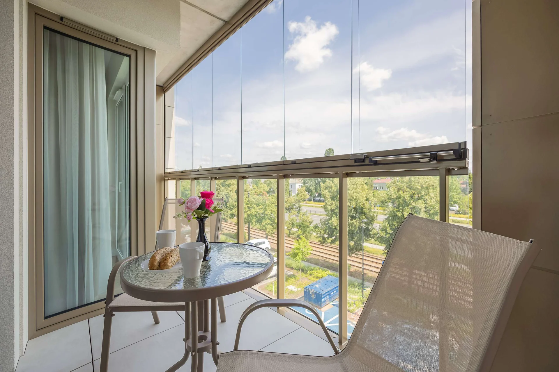 A glass-enclosed balcony is furnished with a round metal table and chairs. The elevated viewpoint looks down onto a green park filled with trees.