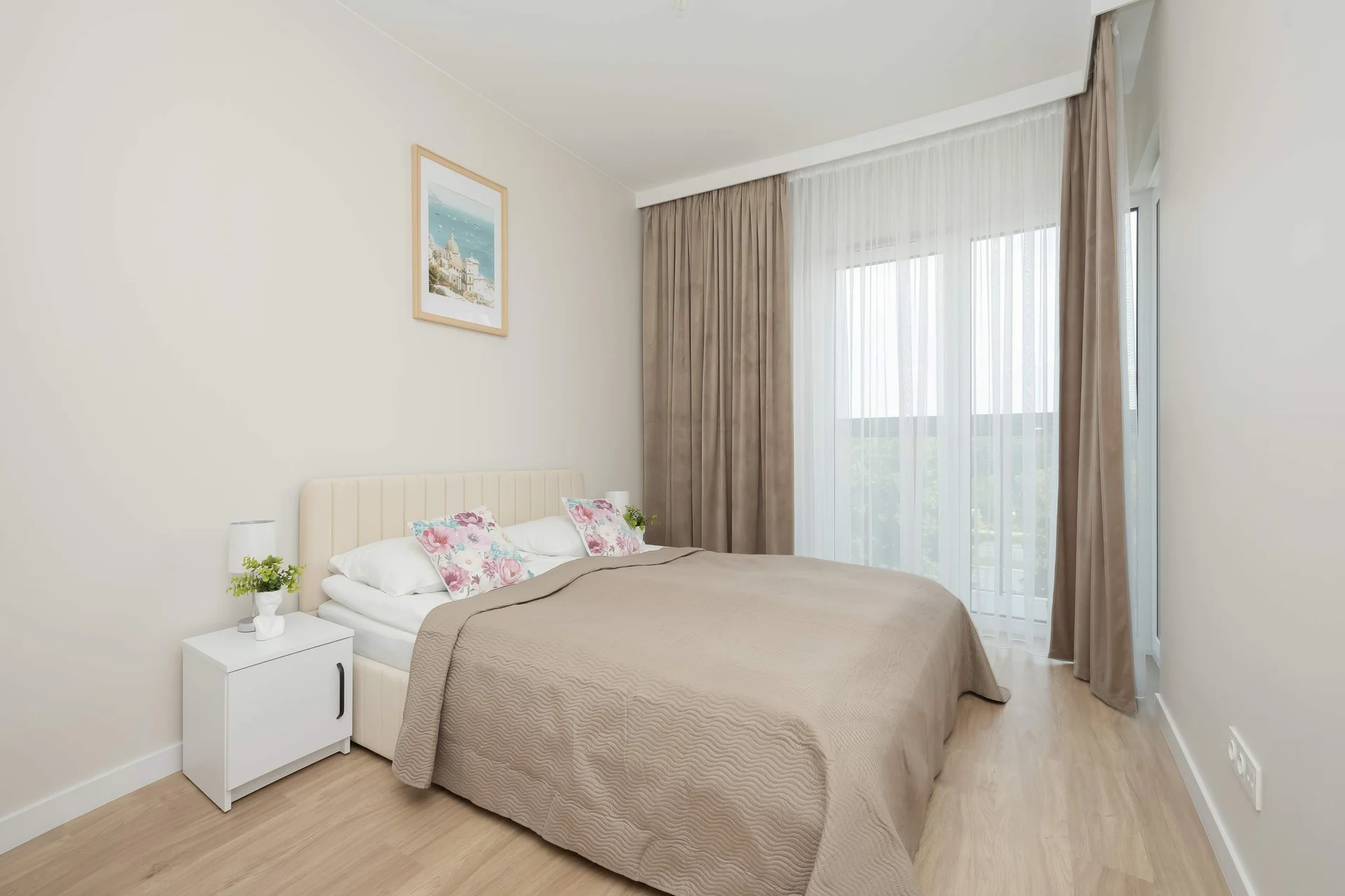 The bedroom displays a double bed styled with a neutral beige coverlet and pillows. A large window with sheer white curtains floods the room with light.