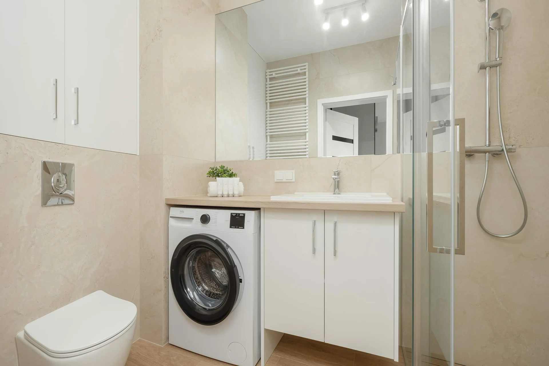 A compact bathroom features a washing machine integrated next to a white vanity. Beige wall tiles surround the space near the toilet and shower.