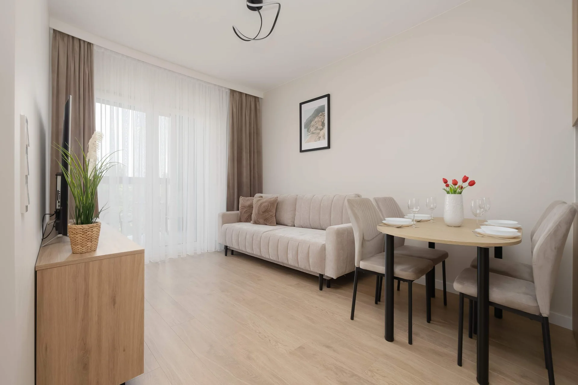 A bright living room features a beige sofa and a small round wooden dining table. Glass doors at the back lead out onto a sunny balcony.