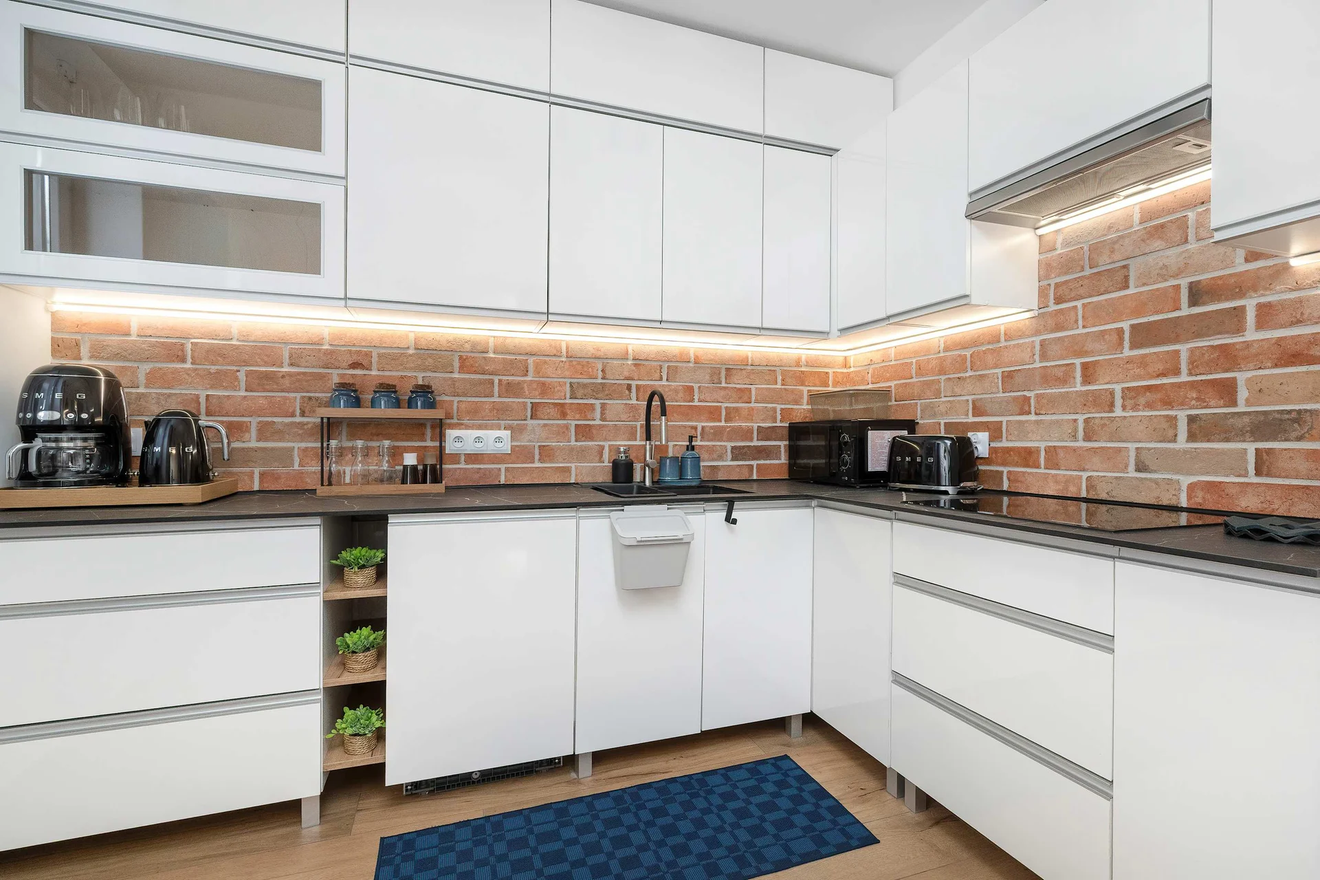 A modern L-shaped kitchen showcases white cabinetry paired with a detailed brick backsplash and under-cabinet lighting. Black appliances and a dark countertop create a sharp, stylish contrast.