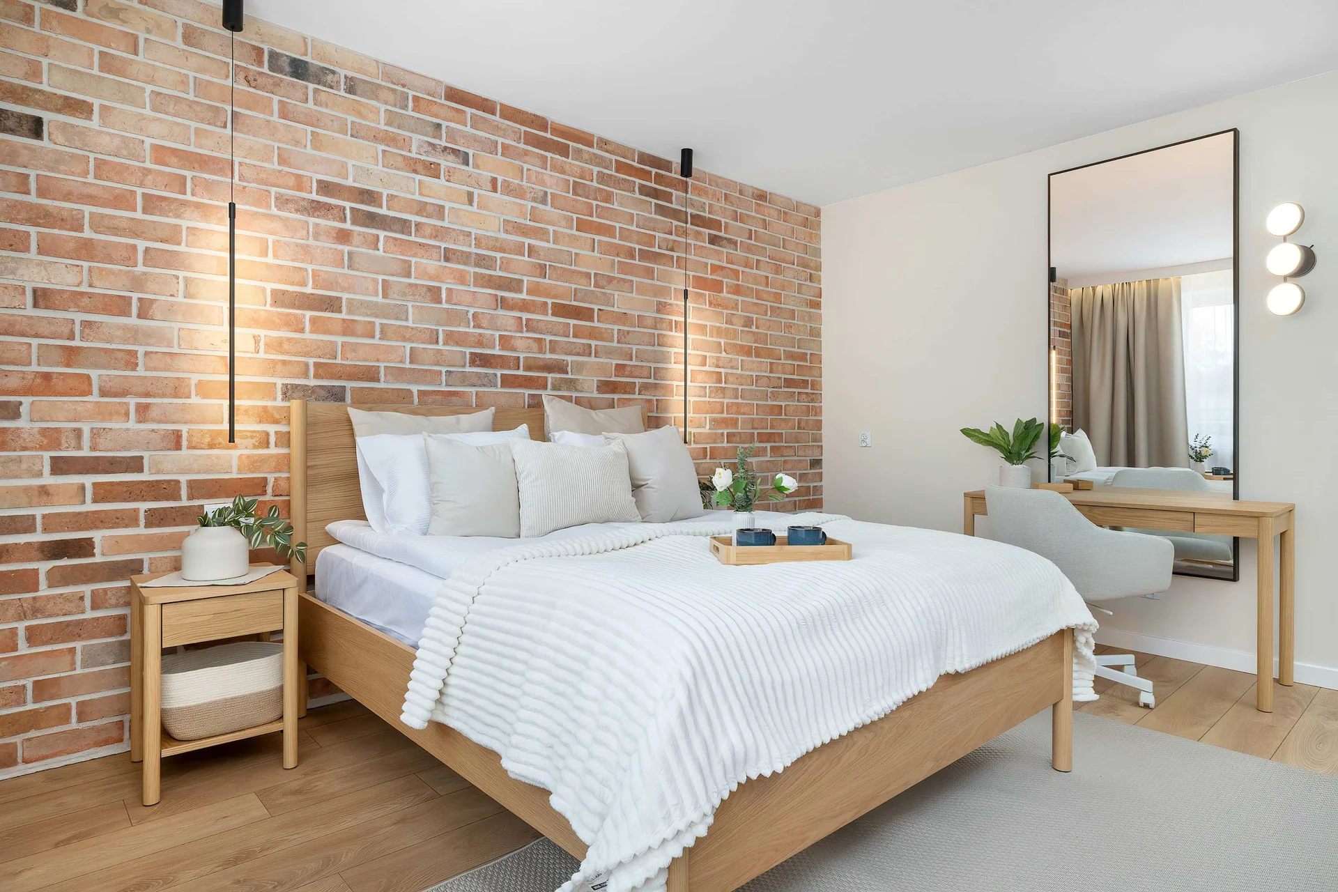 A cozy bedroom features a double bed with crisp white linens and a wooden frame set against a warm brick accent wall. A sleek vanity desk with a circular mirror stands to the right, adding functionality to the modern space.