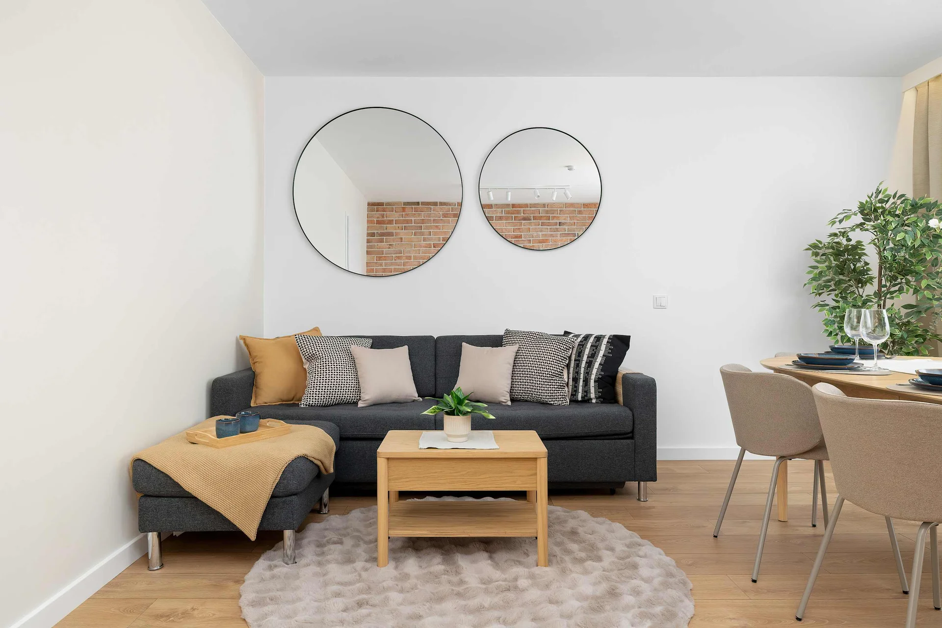An elegant living room layout presents a dark gray sofa adorned with decorative pillows alongside a light wood coffee table. Two circular mirrors hang on the plain wall behind it, reflecting the open-plan dining area.