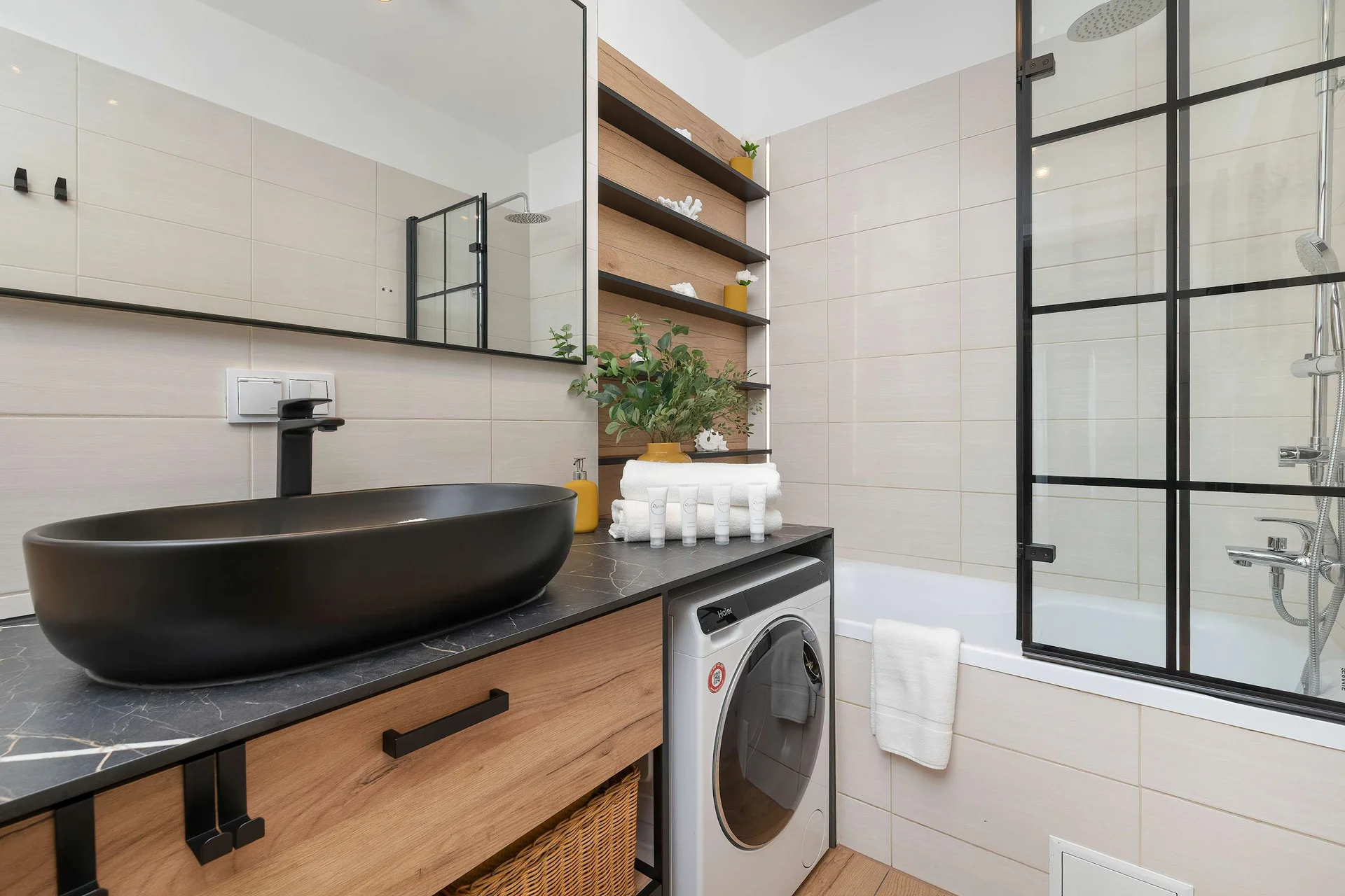 A compact bathroom incorporates a unique matte black vessel sink resting on a wooden vanity. A washing machine is neatly tucked underneath, while a glass shower partition frames the space.