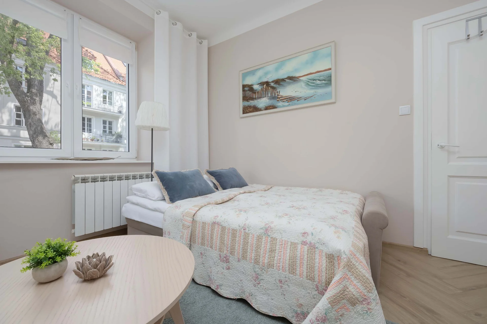 The bedroom section features a neatly made sofa bed adorned with a textured patterned coverlet. A light-colored wooden coffee table sits in the foreground, adding a functional touch to the bedside space.
