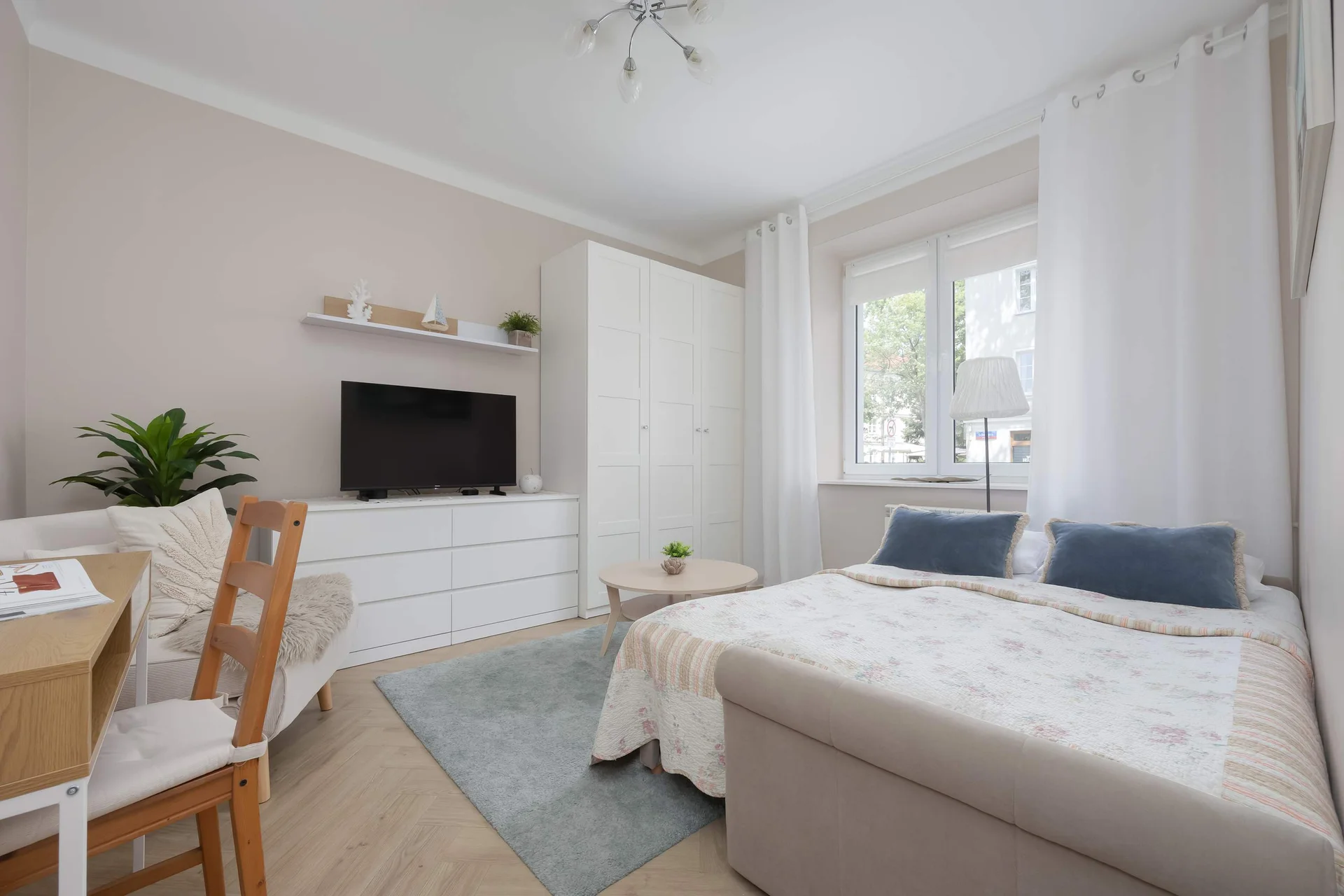 A well-lit studio apartment layout reveals a cozy living area combined with a comfortable sofa bed. A sleek white media console stands against the wall beneath a television, while a small dining table is partially visible on the left.