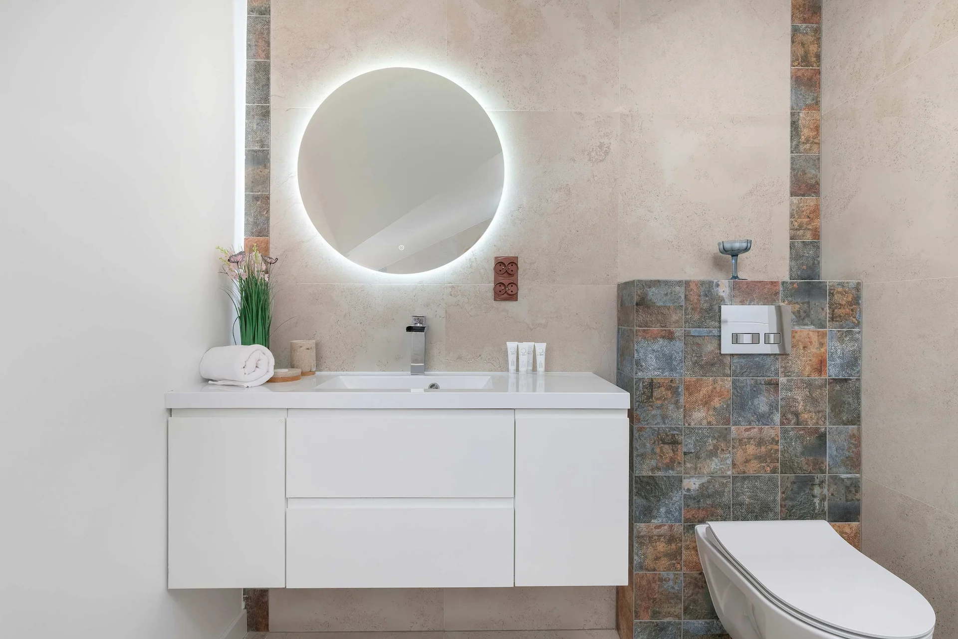 A modern bathroom showcases a floating white vanity with a washbasin below a round, backlit mirror. Patterned mosaic tiles accent the toilet wall.