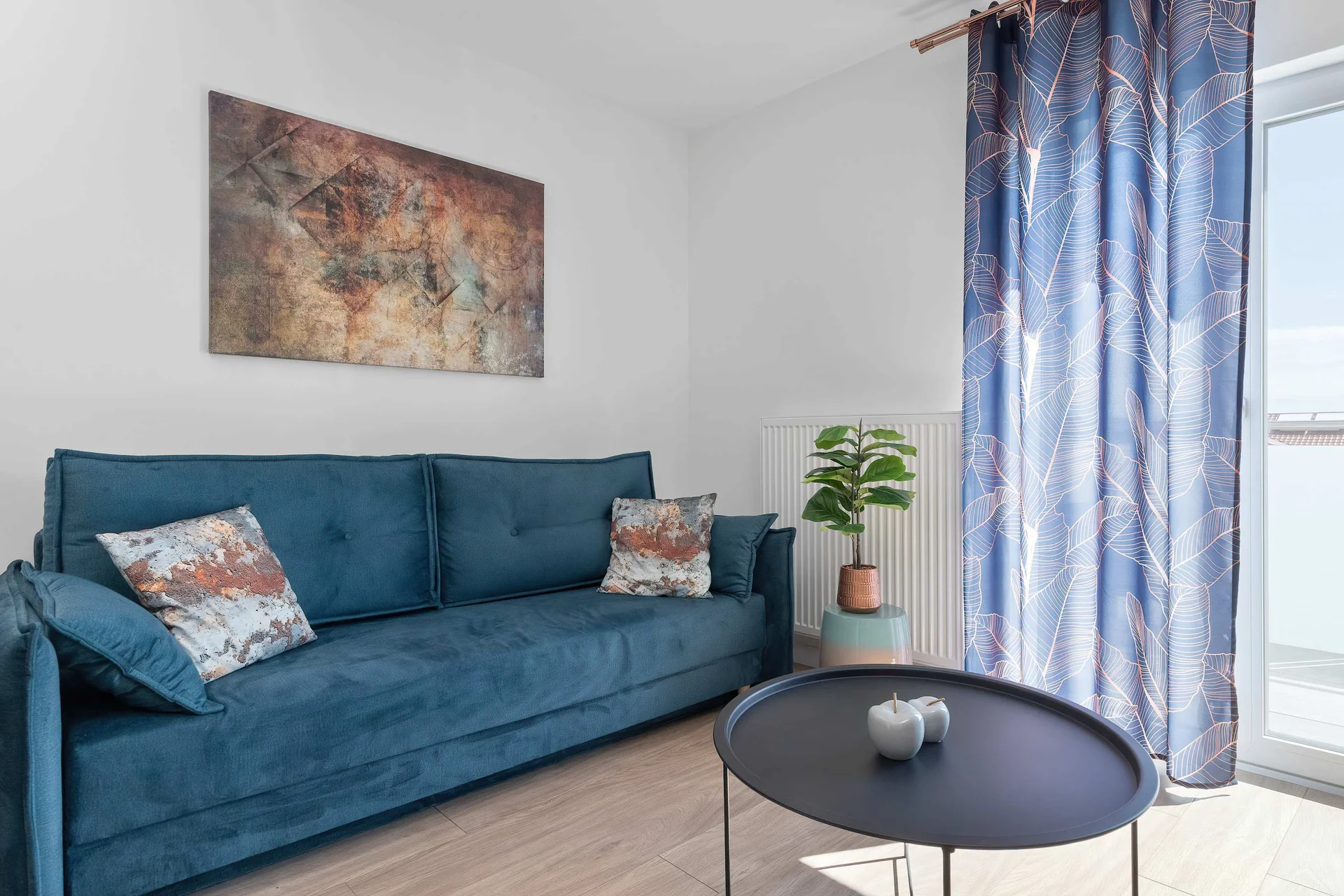 A vibrant teal-blue sofa sits on light wood flooring next to a black circular coffee table. Patterned curtains frame the balcony window in the corner.