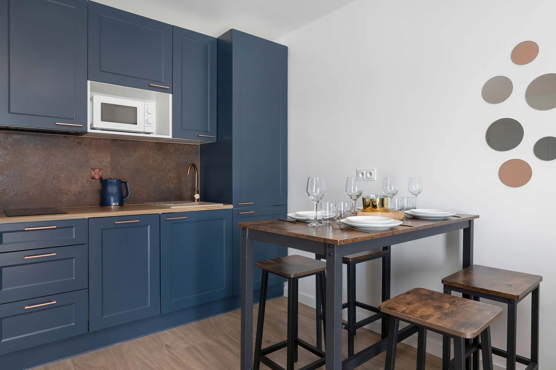 A sleek kitchenette is designed with navy-blue cabinets and a wooden countertop. A high dining table with matching bar stools sits adjacent to it.