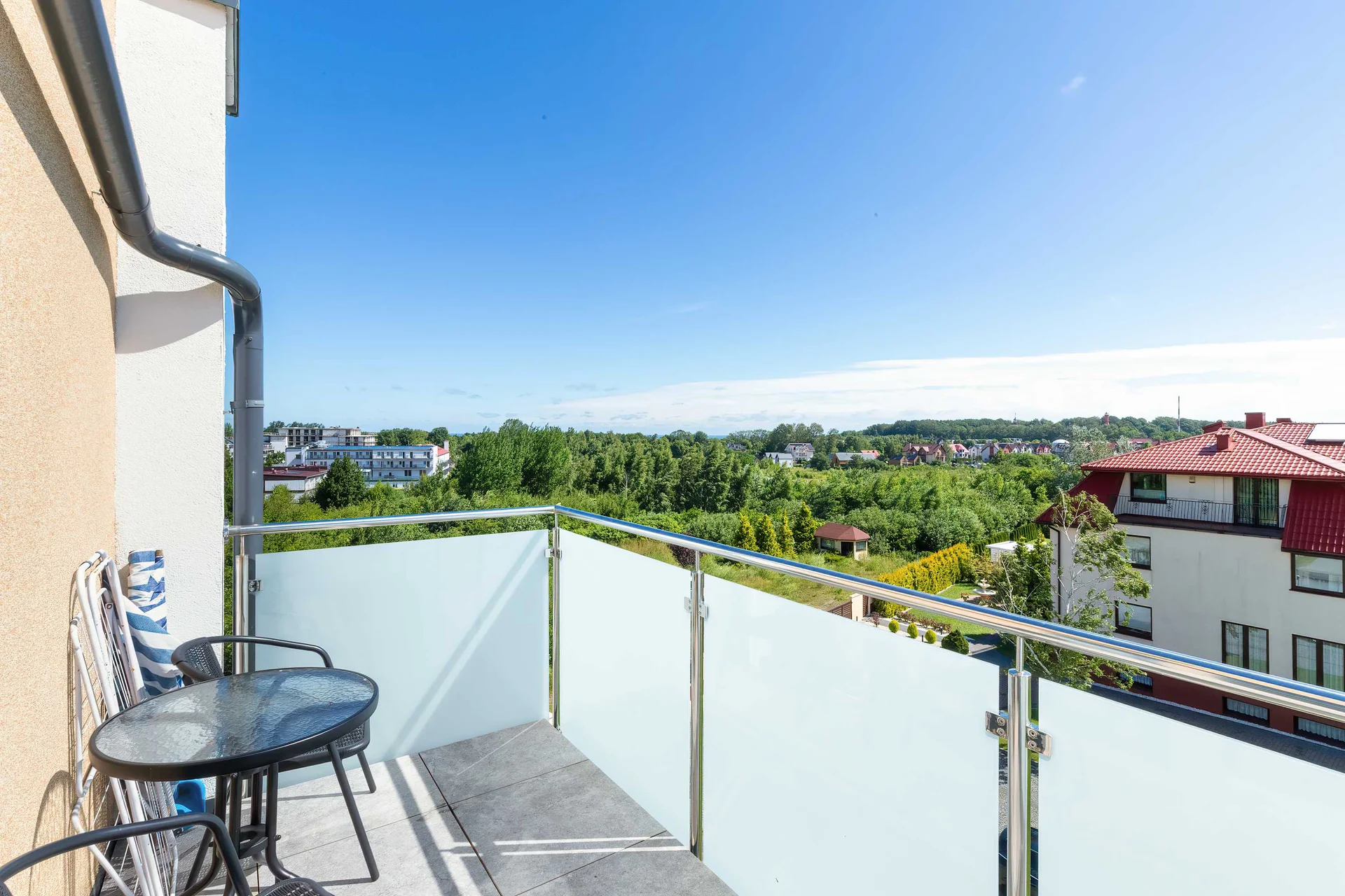 A glass-paneled balcony is furnished with a small patio table and two woven chairs. The elevated viewpoint looks over a lush green horizon.