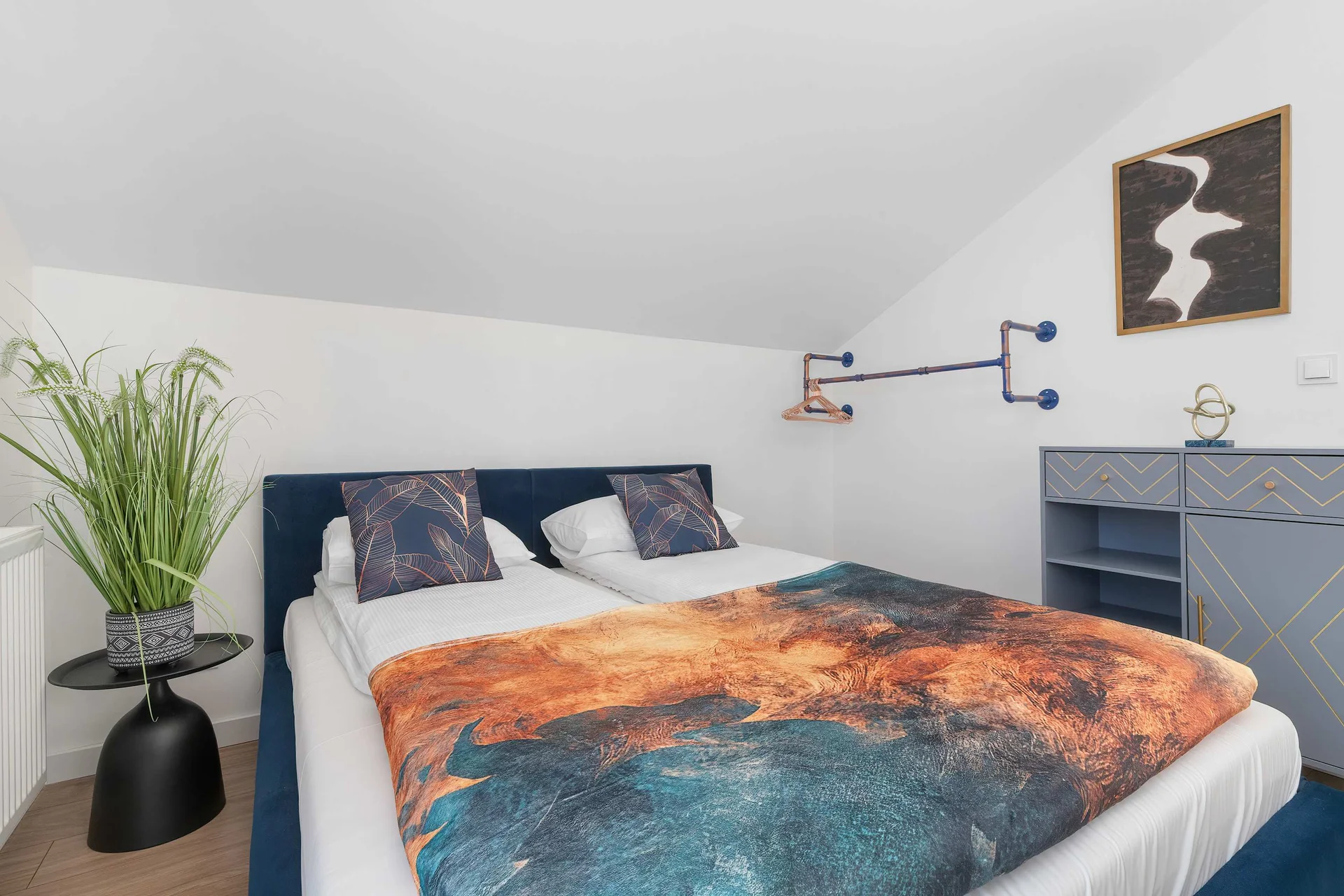 The attic bedroom features a double bed with a colorful, abstract patterned duvet. A potted green plant sits on a black table next to the bed frame.