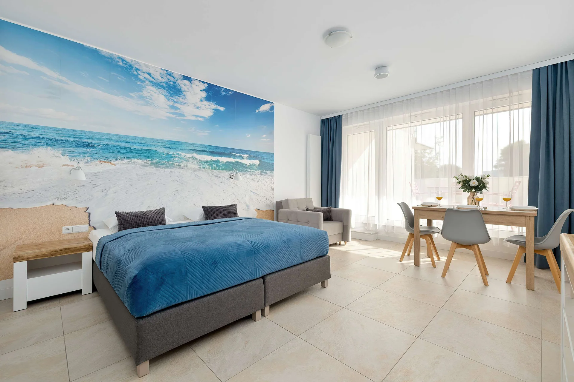 A comfortable bedroom area with a large bed, ocean-inspired wall artwork, and a dining corner nearby. The bright colors and natural light create a peaceful holiday setting.