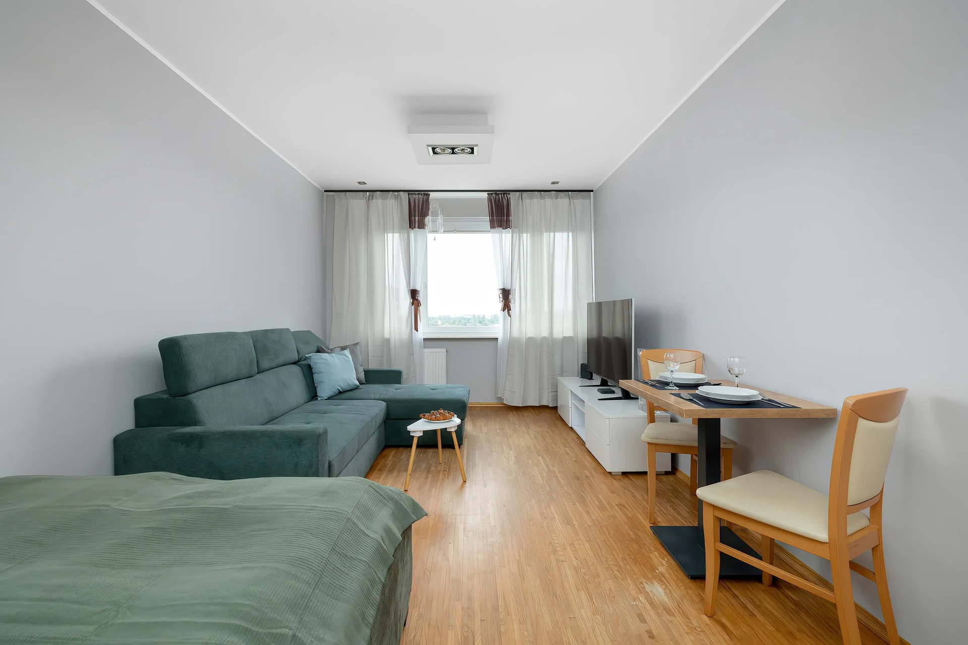 A bright studio apartment with a double bed, a dining table, and large windows. The space is simple, modern, and comfortable.