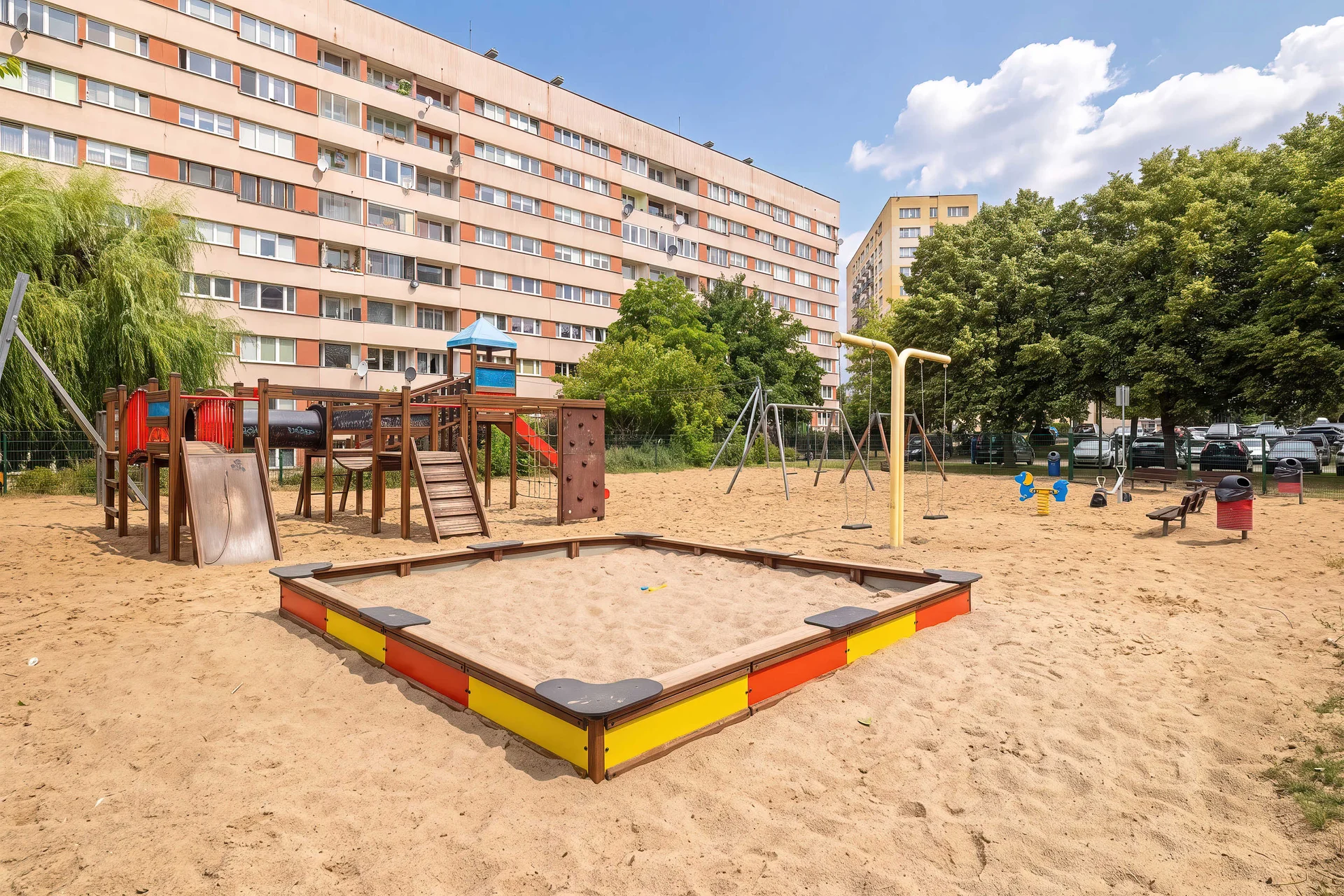 A children's playground with a sandbox and play equipment. The area is located next to a residential building.