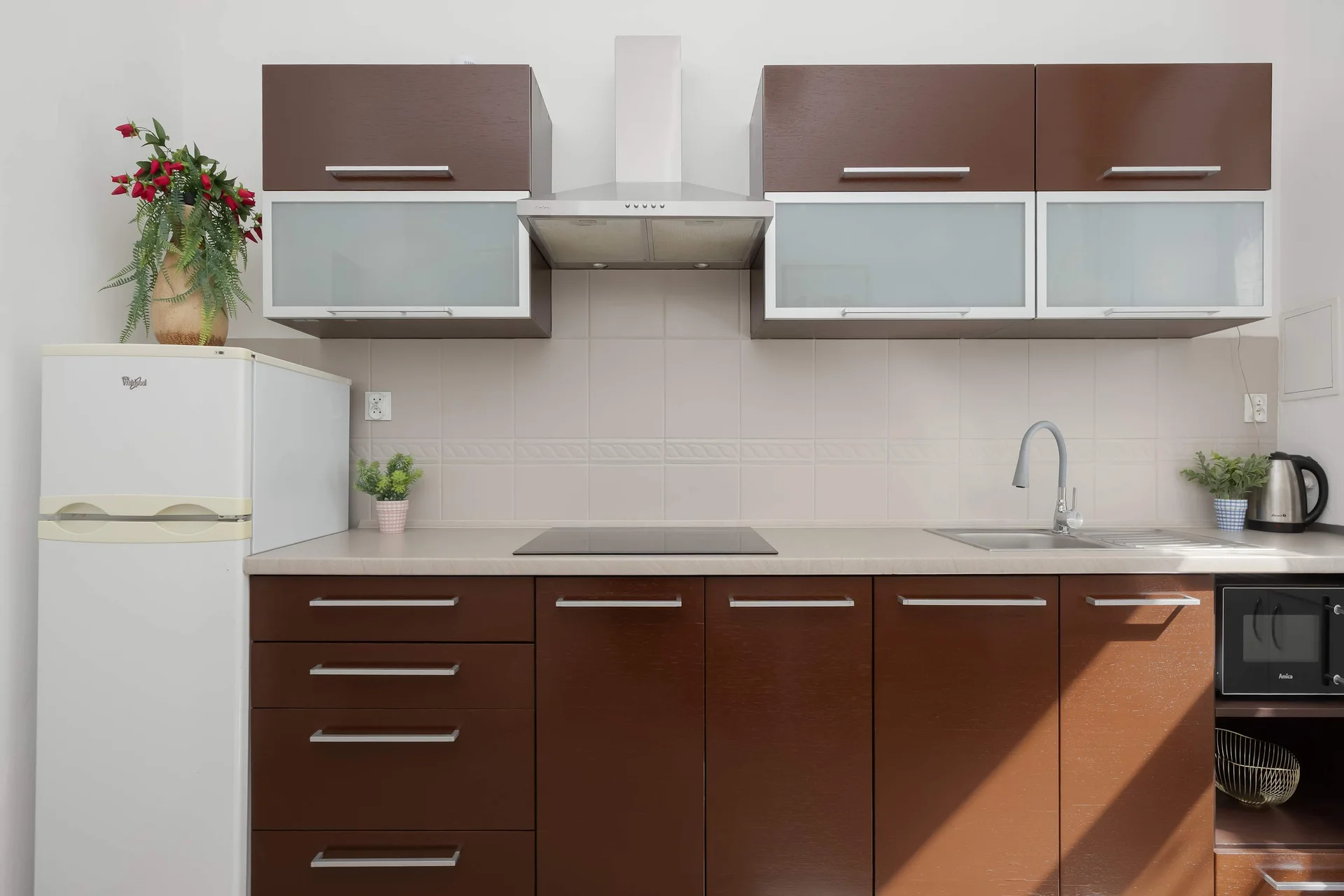 A modern kitchen setup showcases dark brown lower cabinetry and frosted glass upper cabinets. The countertop includes an integrated cooktop and a stainless steel sink with a high-neck faucet. A white refrigerator stands on the left side of the kitche