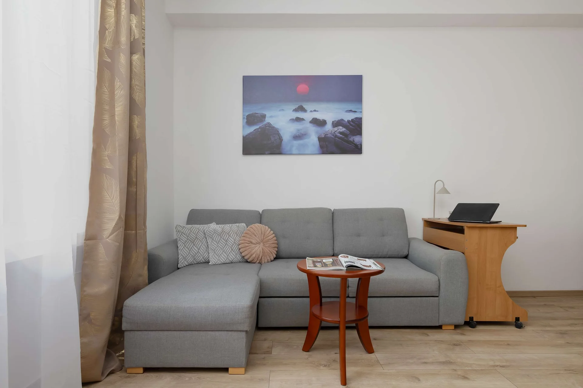 A cozy living room area features a grey sectional sofa adorned with decorative pillows. A small wooden coffee table sits in front of the couch, and a computer desk is visible to the right. A landscape painting hangs on the wall above the seating setu