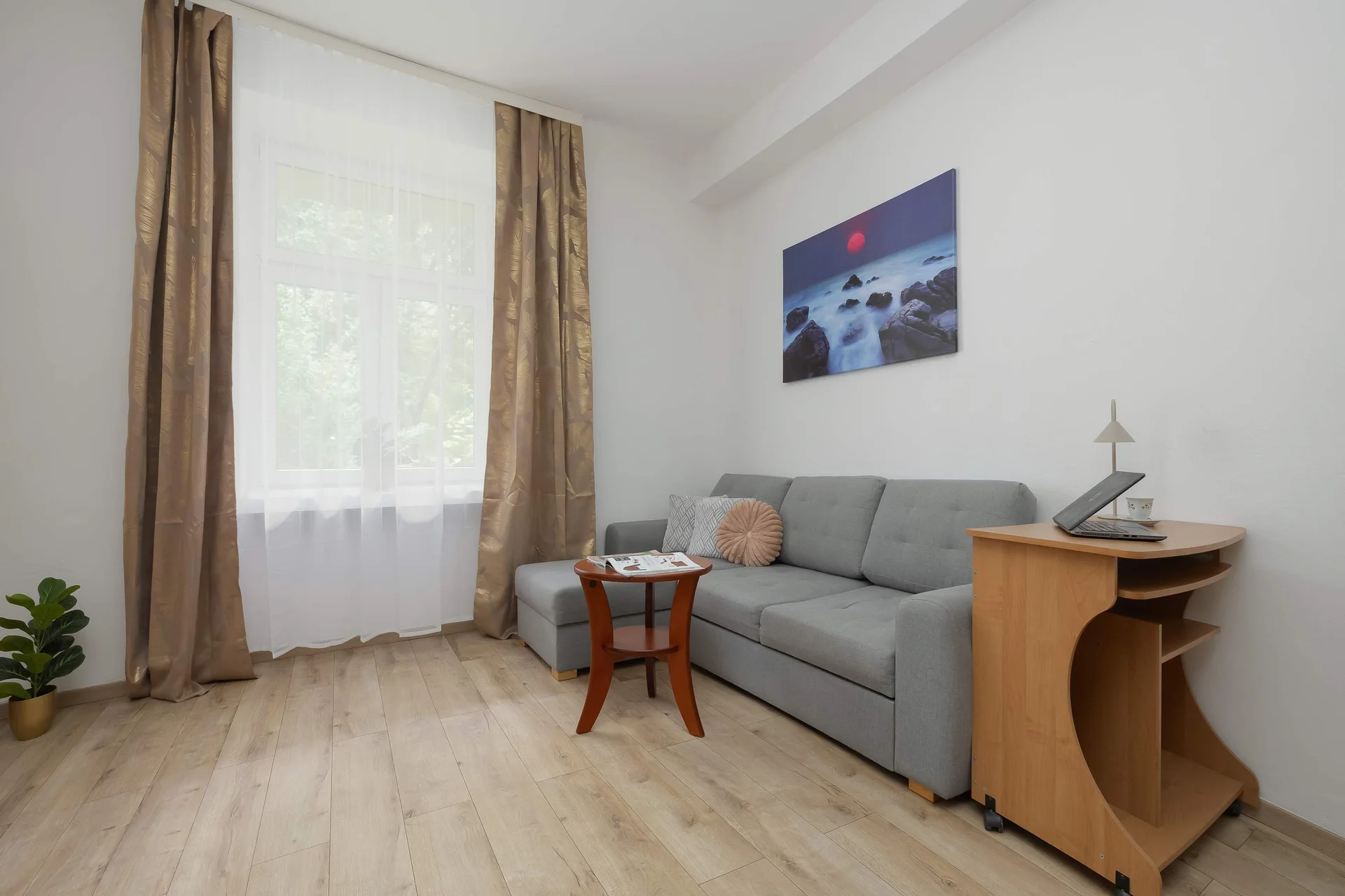 A bright living space is shown from an angle highlighting a large window dressed with beige curtains. The grey sectional sofa and a small coffee table occupy the center of the room.