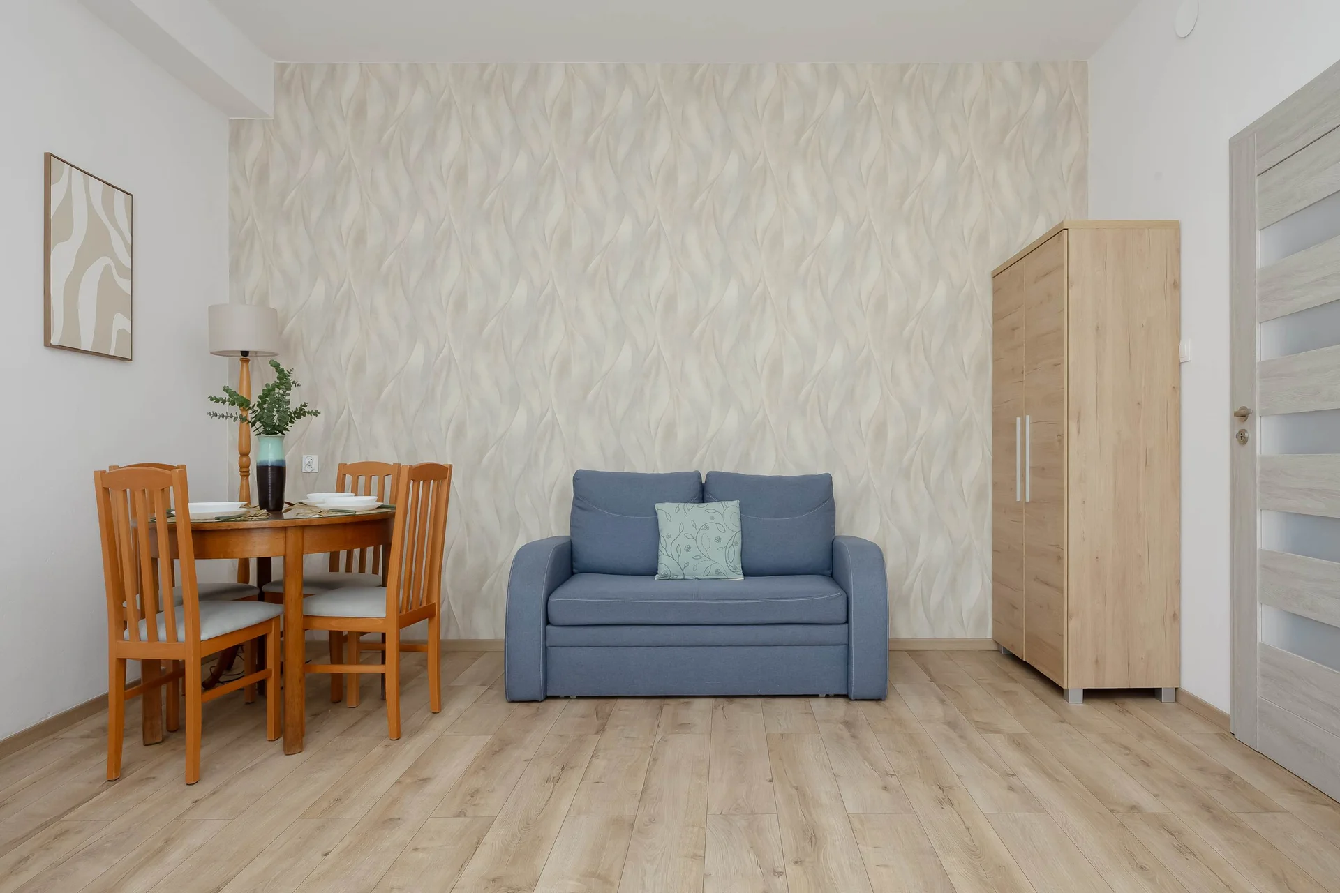 An open-concept room contains a wooden dining table paired with matching chairs on the left. In the background, a blue sofa rests against a patterned wall next to a tall wooden wardrobe. Light-colored laminate flooring extends across the entire space