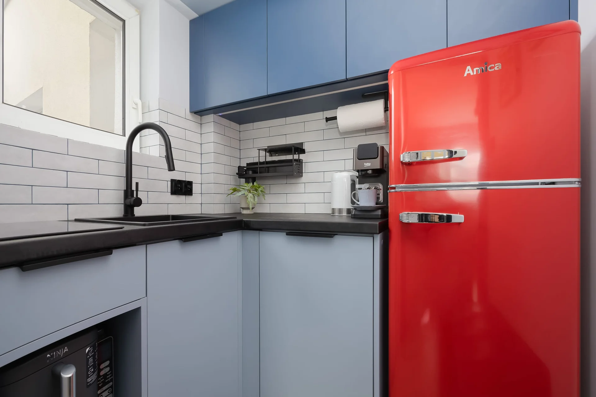Modern kitchenette with blue cabinets, a retro-style red refrigerator, subway tile backsplash, and all the essentials for preparing meals.
