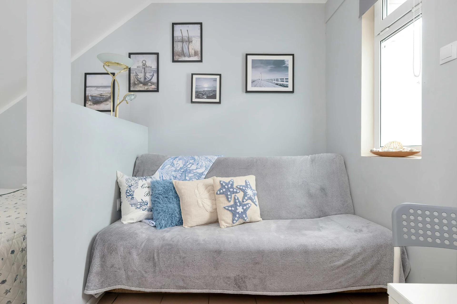 A grey upholstered sofa bed is neatly tucked into a corner, adorned with decorative blue and patterned pillows. Small framed pictures hang on the wall above, adding a personal touch to the compact living space.