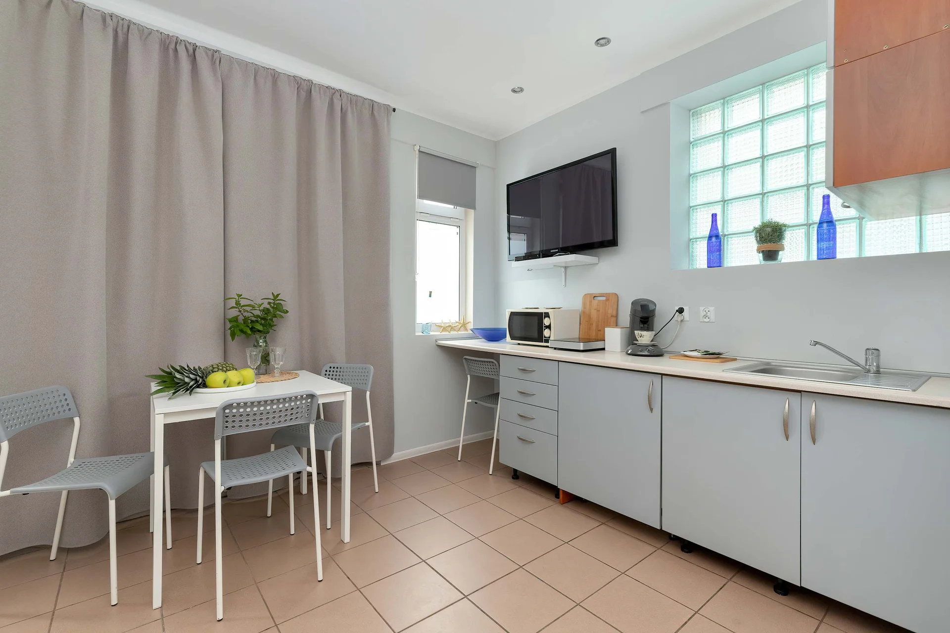 A functional kitchenette and dining area showcase light grey cabinetry topped with a dark countertop and essential appliances. A small white dining table with two chairs sits nearby under a bright window.