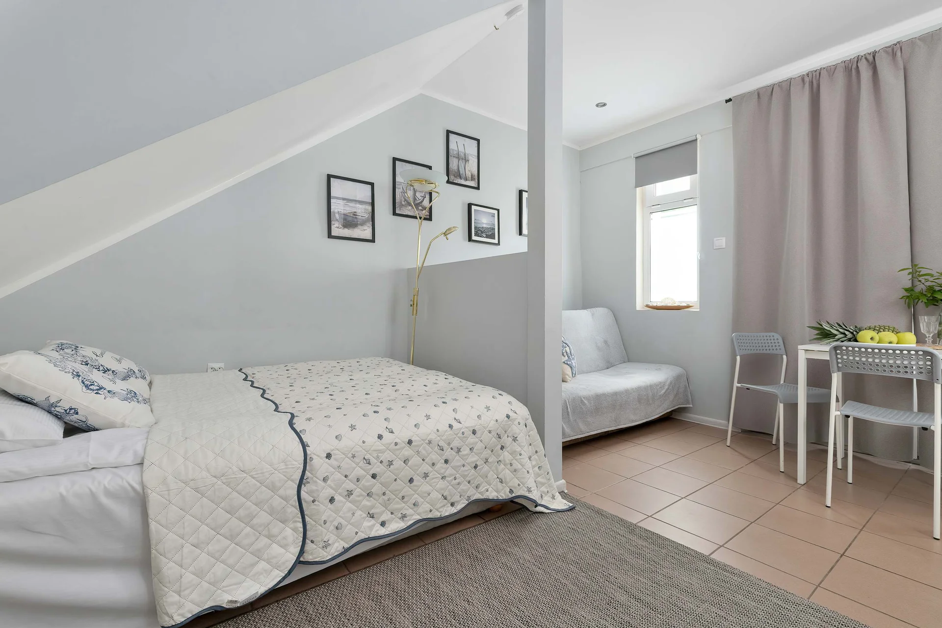 A cozy studio apartment space features a bed with a patterned white coverlet positioned under a slanted ceiling. In the background, a small dining table with chairs and a grey sofa complete the functional living area.