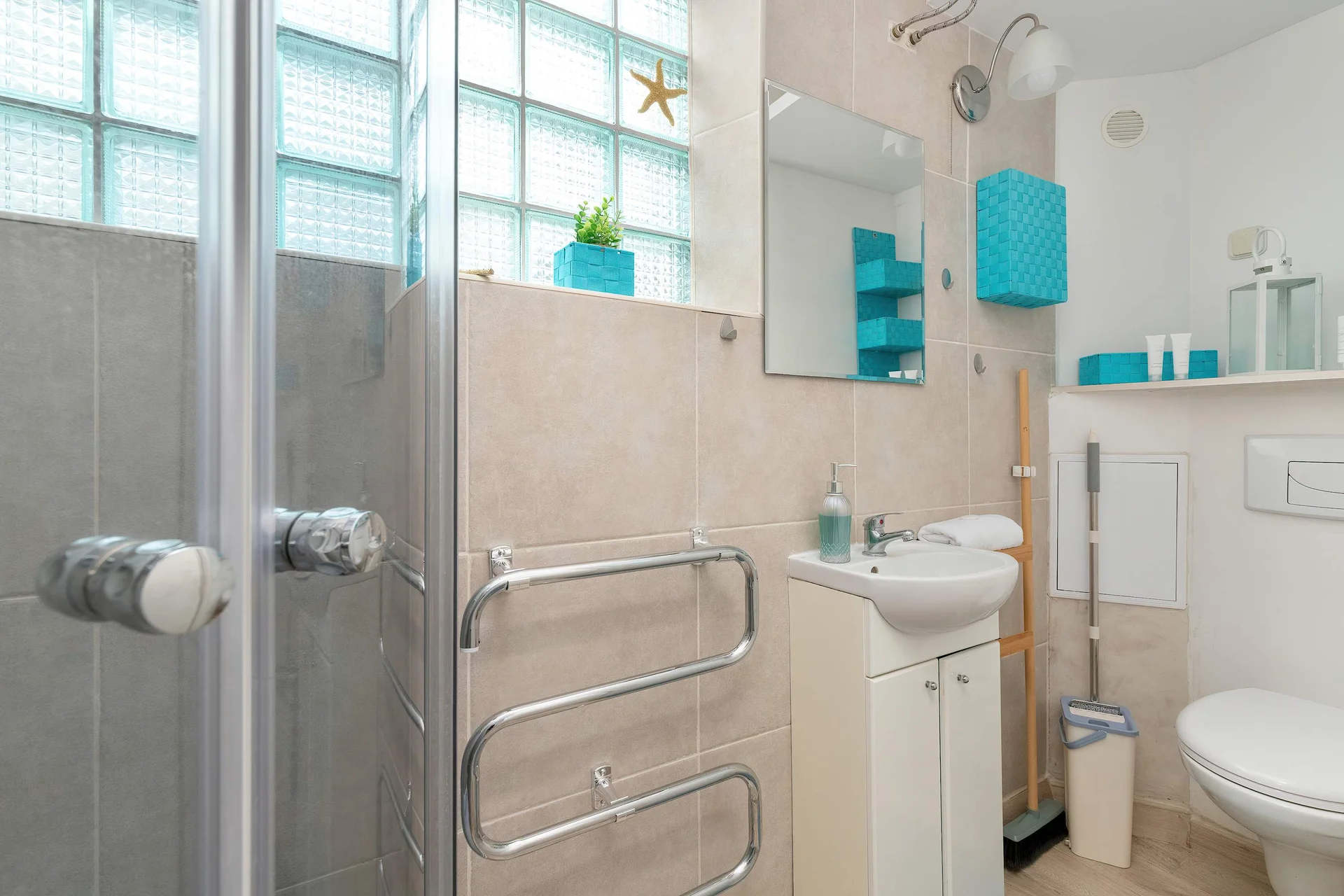 A compact bathroom includes a white vanity sink setup with a mirror hung on the tiled wall. A heated towel rail and a glimpse of the toilet fill out the rest of the functional space.