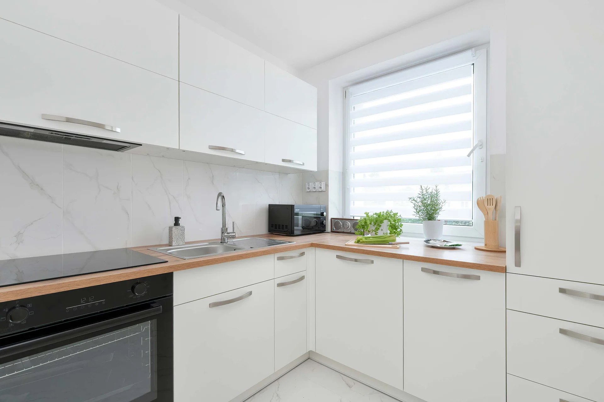 A modern white kitchen offers sleek cabinetry, built-in appliances, and generous countertop space. A large window above the sink fills the room with daylight.