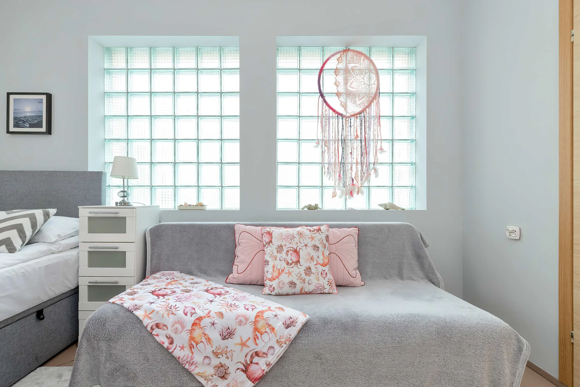 A bedroom area centered around a bed with gray bedding, accompanied by a smaller sofa with patterned pillows. Glass block windows allow natural light to filter into the space.