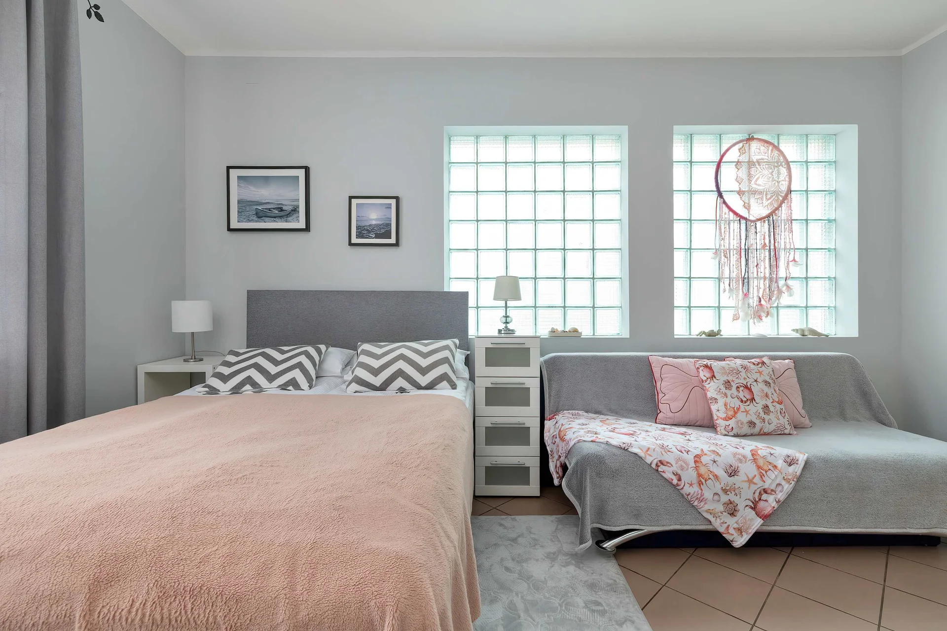 A sleeping area furnished with a bed dressed in light-colored linens and an adjacent sofa with decorative cushions. The room is accented by glass block windows and neutral-toned decor.