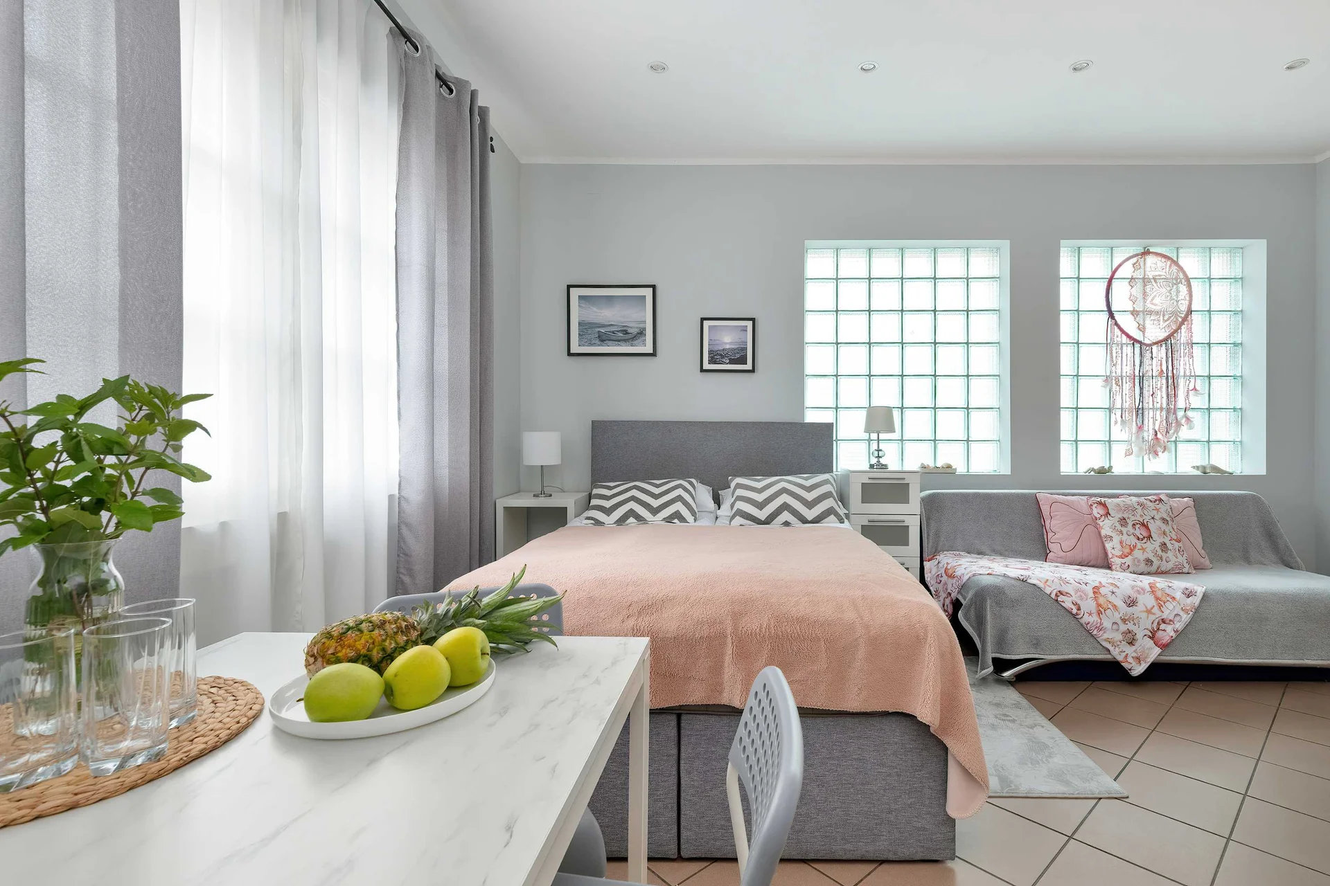 A studio room featuring a double bed with a light-colored throw, a dining table with fruit, and a sofa positioned near glass block windows. Soft gray curtains frame the windows, creating a bright and airy atmosphere.