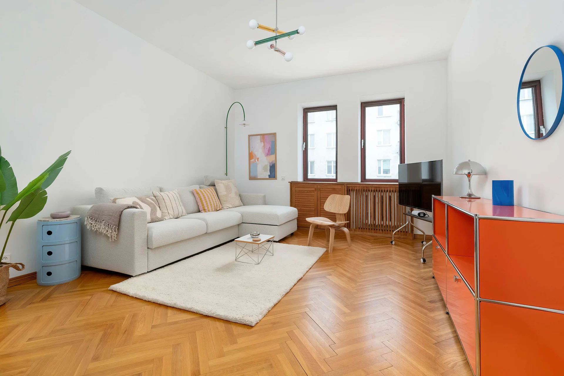 A bright living room with a beige sofa, wooden floor, and large windows creates a cozy atmosphere. Colorful accents add a modern touch to the space.