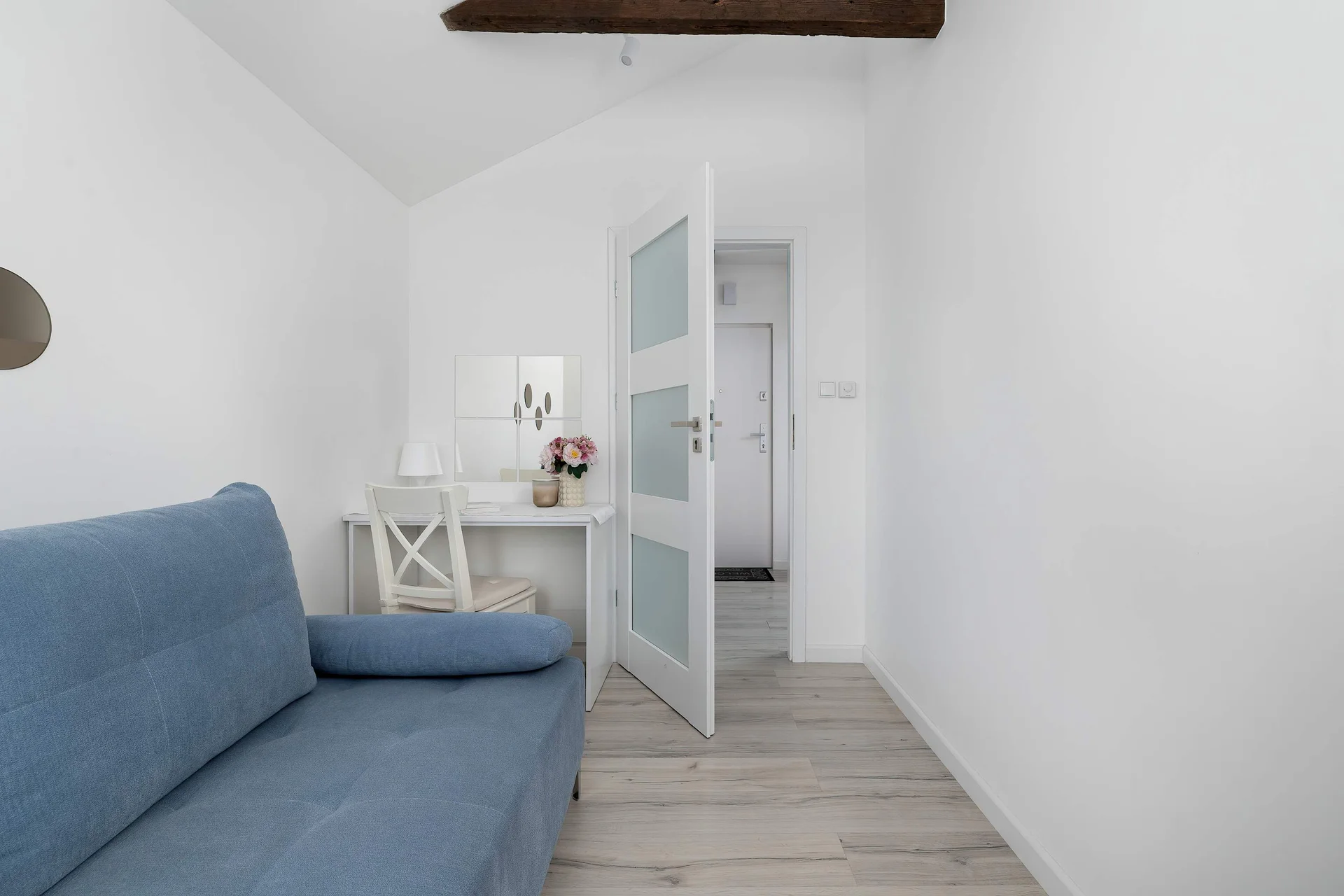 A blue sofa is placed in a bright living area with light wooden flooring. The hallway leads to the rest of the apartment.
