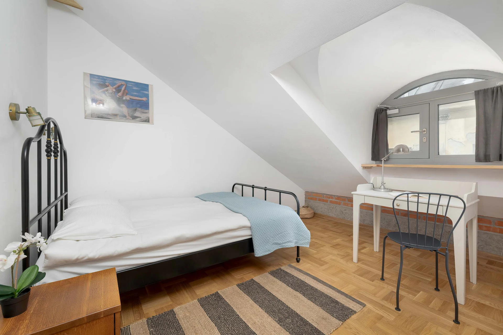 A bright bedroom situated under a sloped ceiling with a single bed dressed in white linens and a light blue throw. Next to the bed is a small wooden nightstand, and a dark study desk with a chair sits near the window.