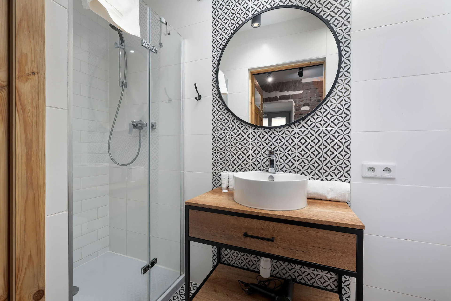 A sleek bathroom focusing on a modern vanity unit with a wooden countertop and a round vessel sink. A large circular mirror with a black frame hangs on a decorative black-and-white patterned tile wall.