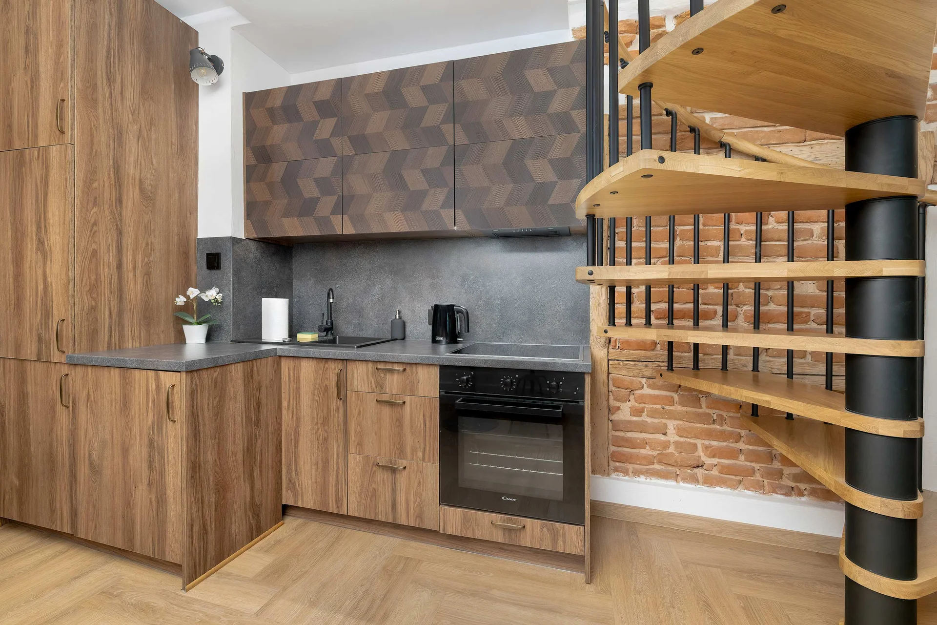 A modern kitchen area showcasing wooden cabinets, a dark countertop, and integrated appliances. A striking spiral staircase with wooden steps and a black metal frame is visible on the right.