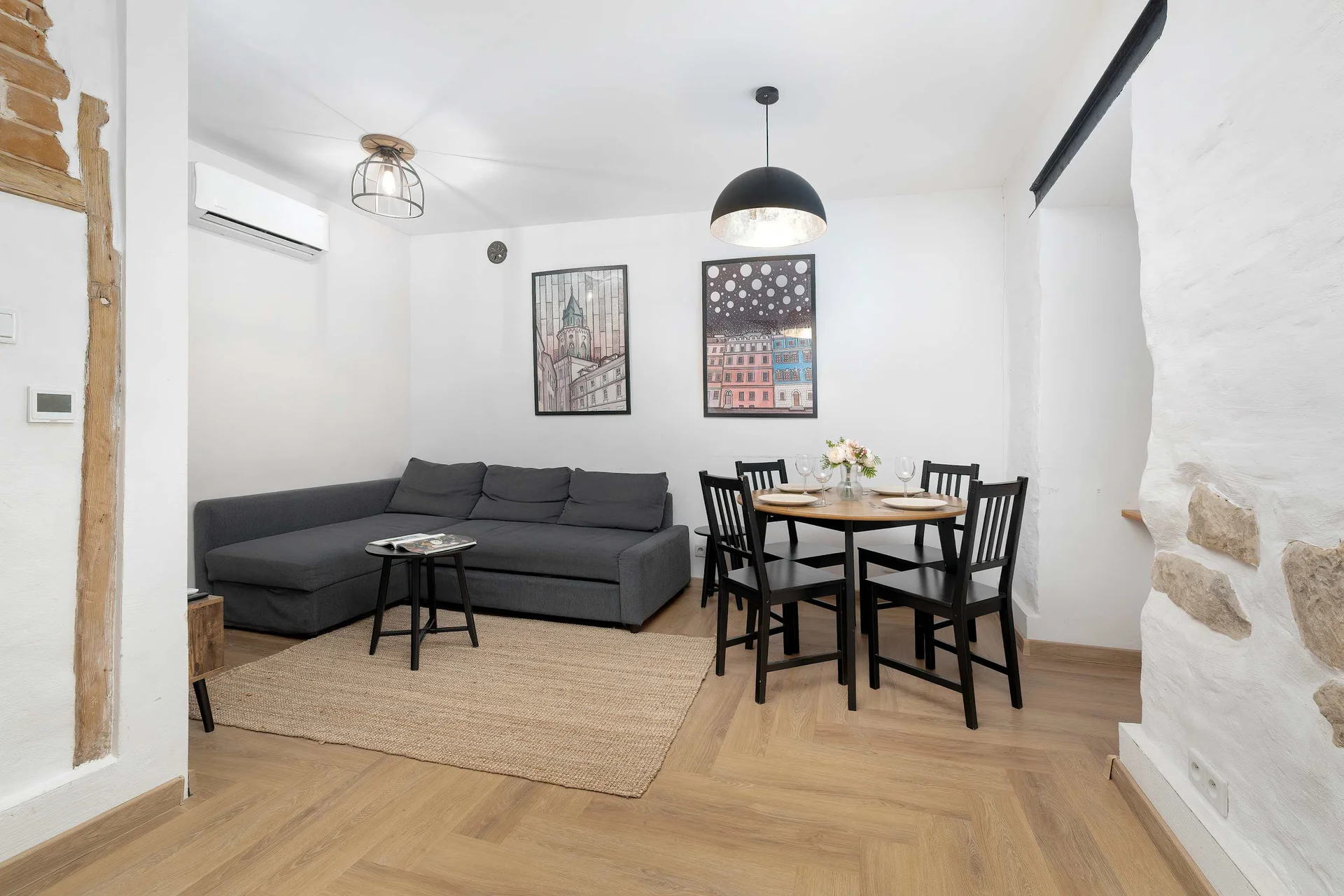 An open-concept living space highlighting a comfortable grey sectional sofa and a round dining table with four chairs. The room features historic elements like an exposed stone wall and warm wooden floors.