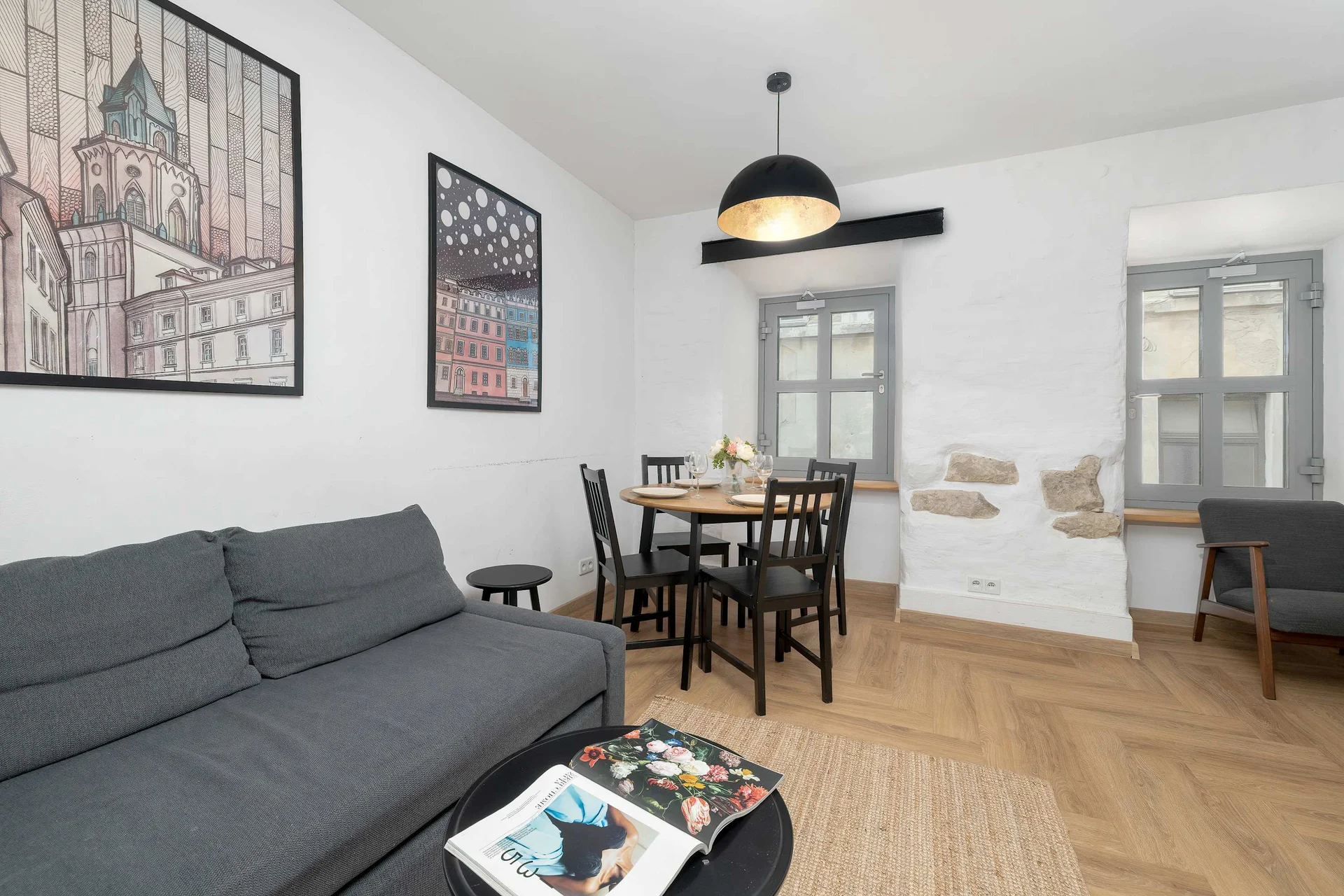 A cozy living area featuring a dark grey sofa and a small dining setup with a table and chairs. Light-colored wooden floors run throughout the space under warm, inviting lighting. Art pieces on the wall and a small coffee table complete the comfortab
