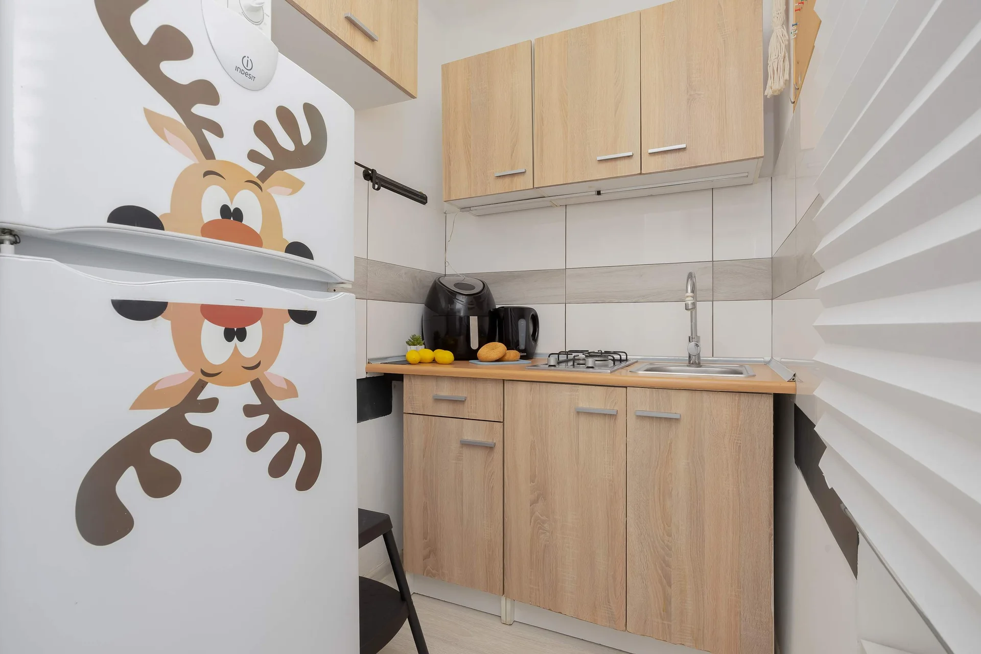 A compact kitchen corner displays light wood cabinetry paired with a clean countertop and sink. A white refrigerator stands on the left, playfully decorated with a festive deer decal.