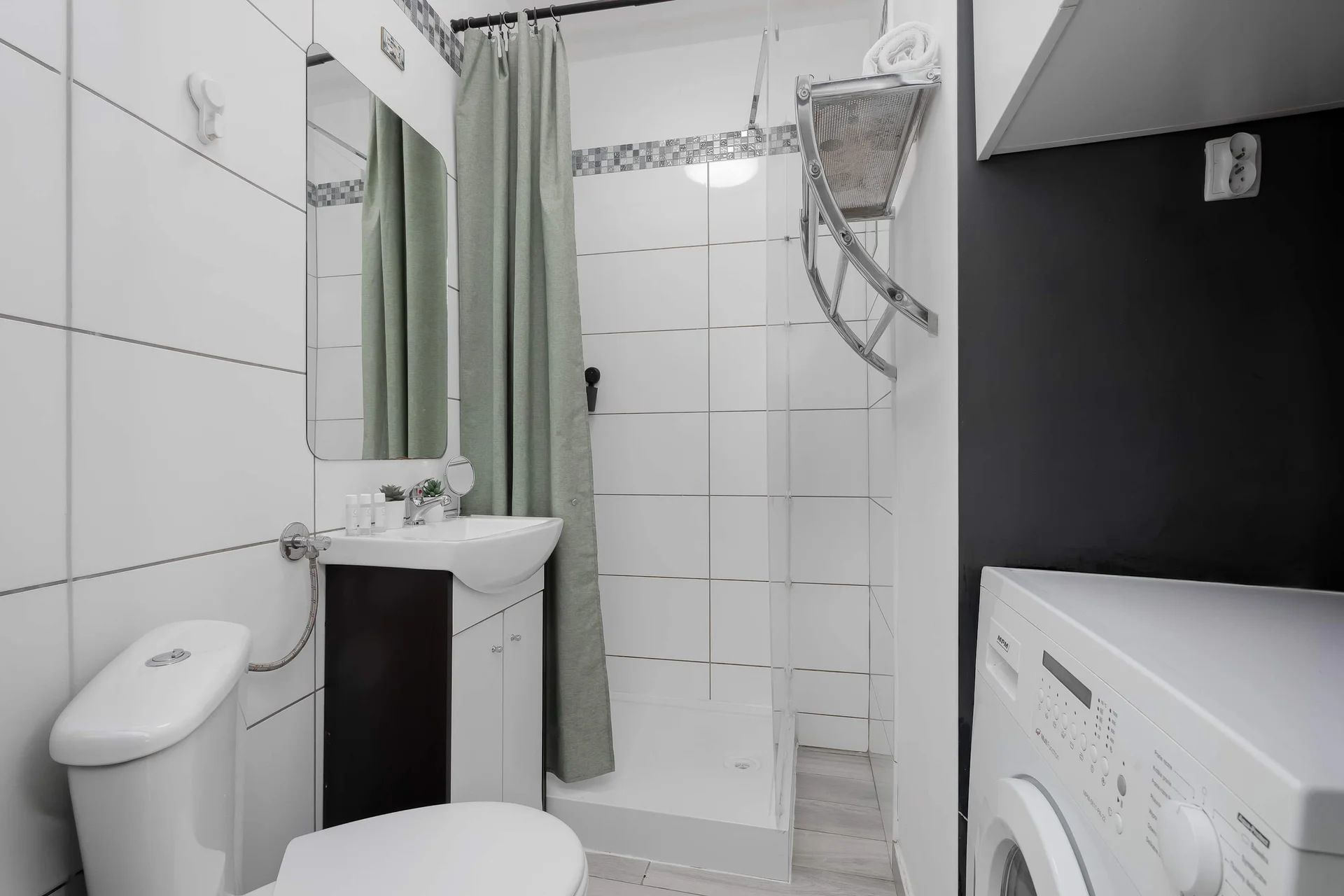 A functional, tiled bathroom contains a white toilet and a compact sink vanity unit. A gray shower curtain hangs ready to enclose the stall in the background. A white washing machine is partially visible on the right.
