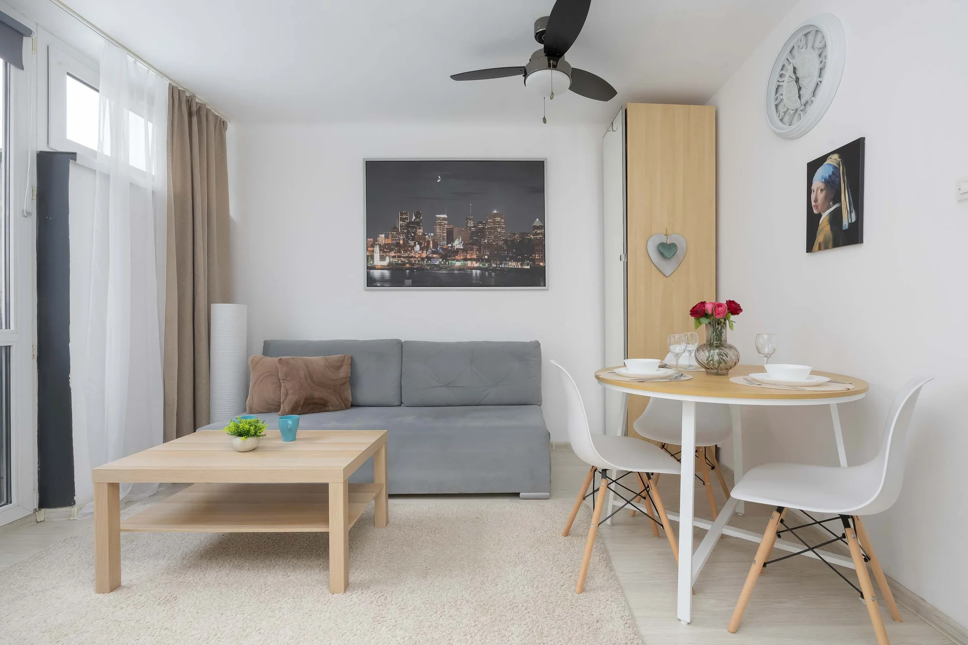 A bright and airy studio apartment layout includes a gray sofa and a light wood coffee table. A round dining table with two white chairs is nestled comfortably in the corner.